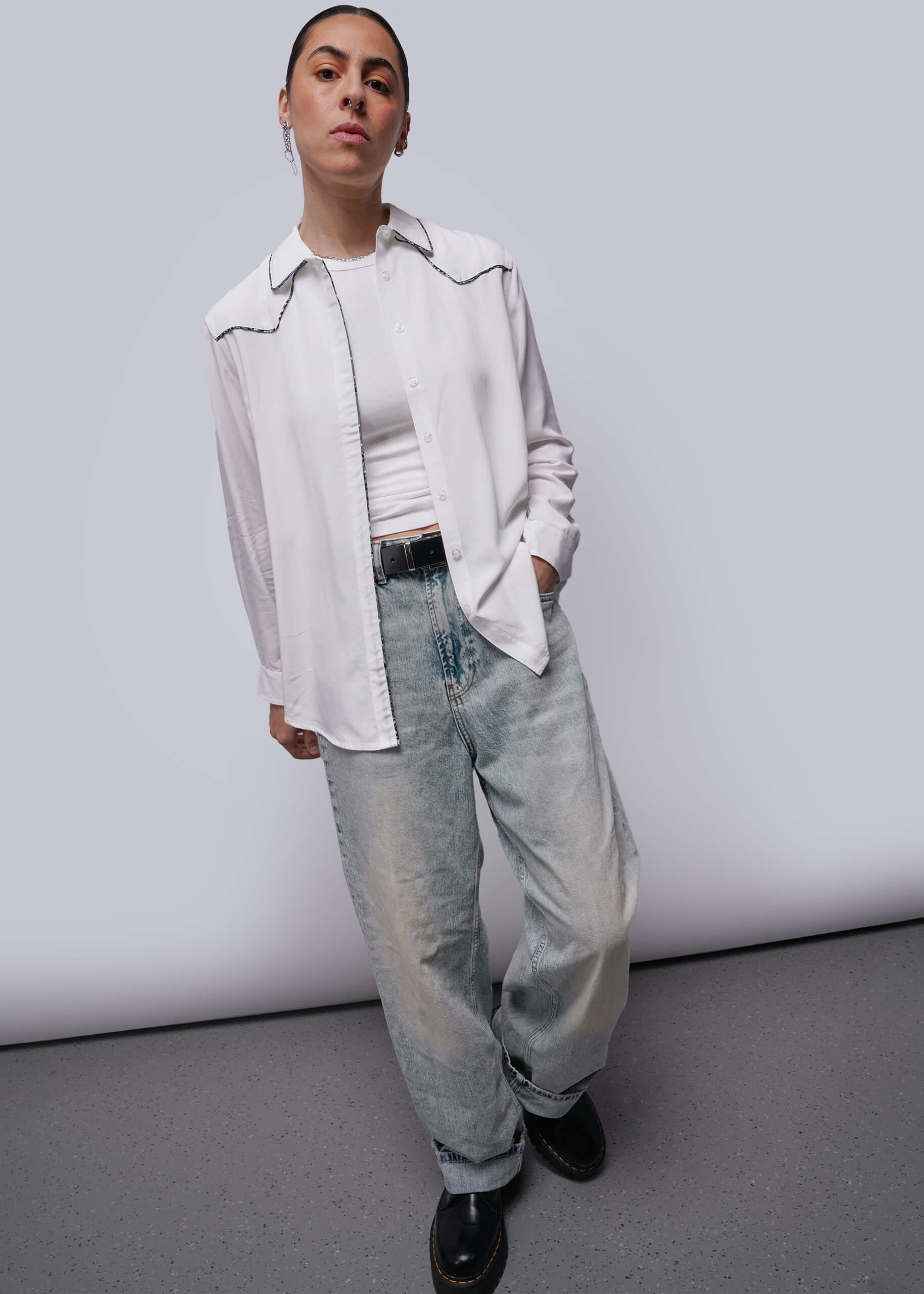 S=Person wearing a white button-up shirt, white top, and light blue jeans against a plain background