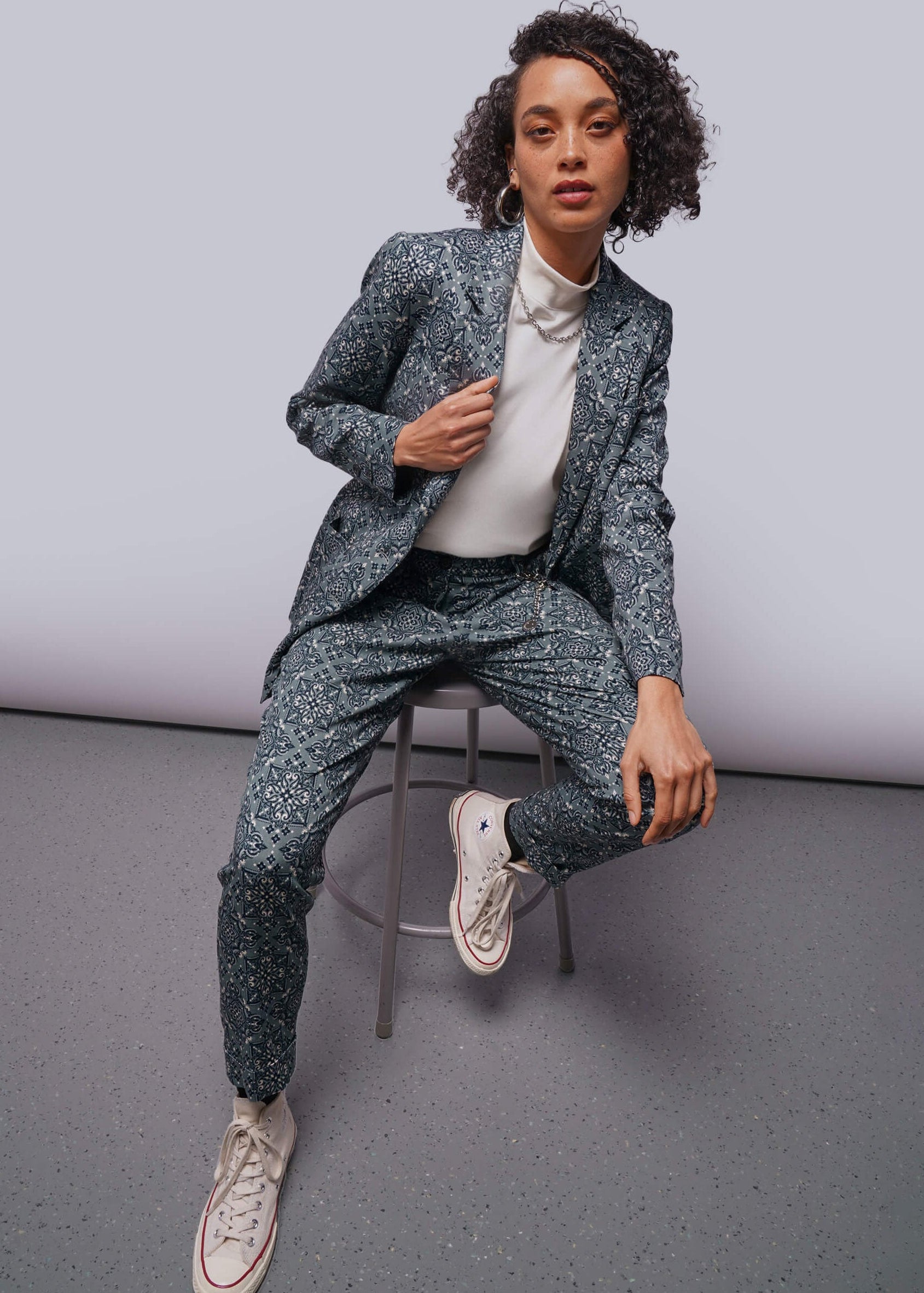 S=Person wearing a patterned suit sitting on a stool against a plain background