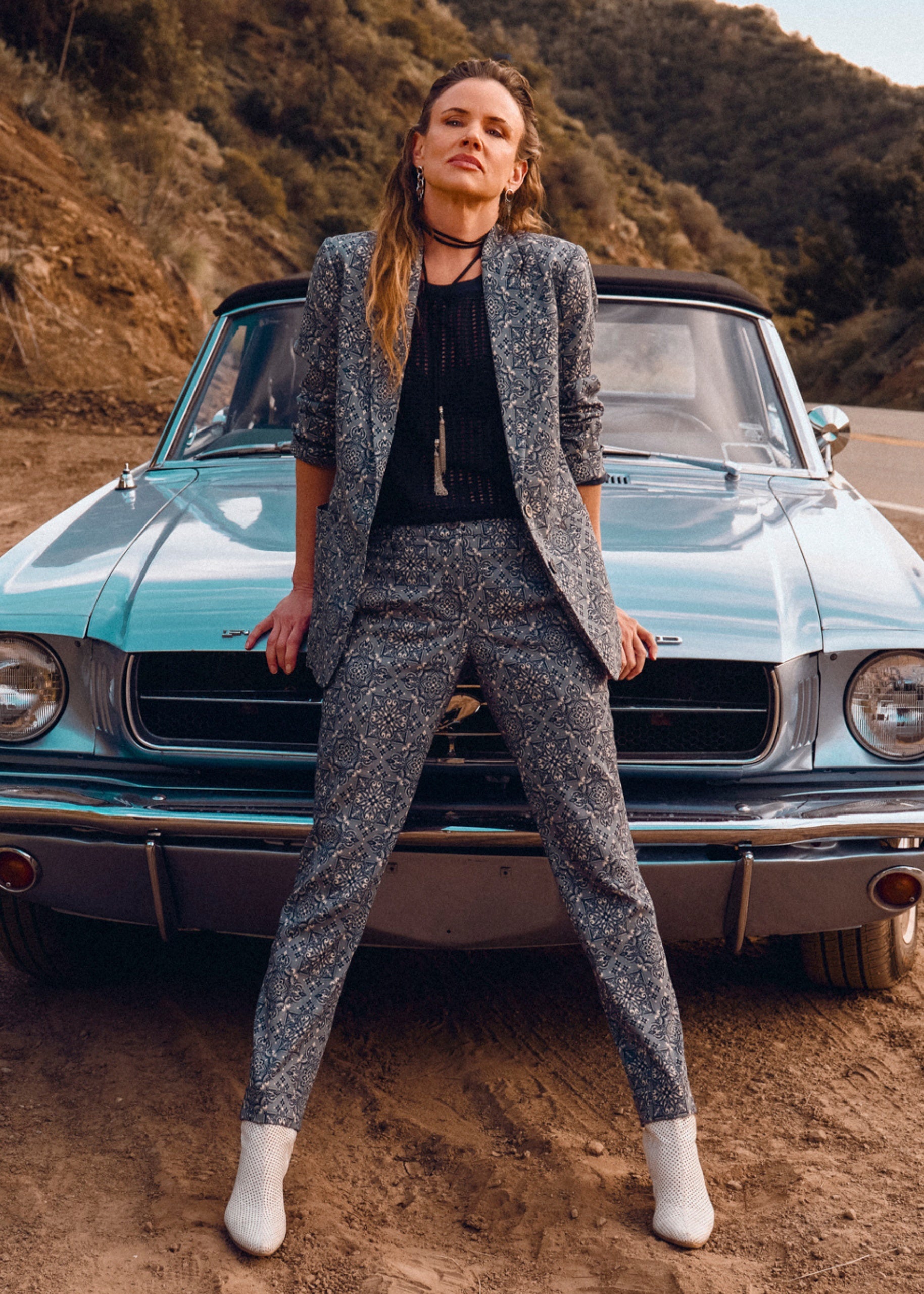 Woman in a patterned outfit standing in front of a vintage car on a dirt road.