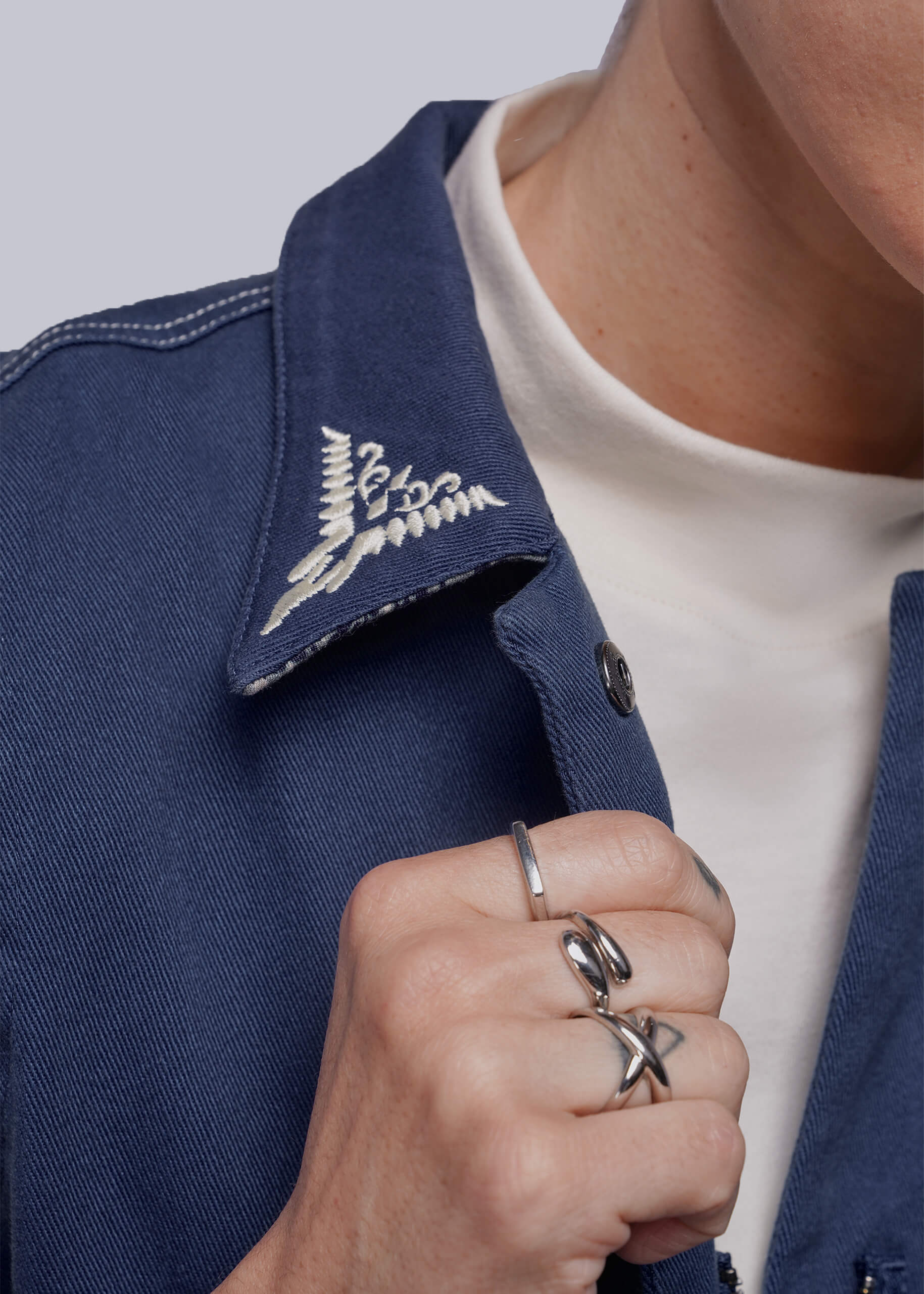 Close-up of a person wearing a blue jumpsuit with collar embroidery, a white shirt, and multiple rings.
