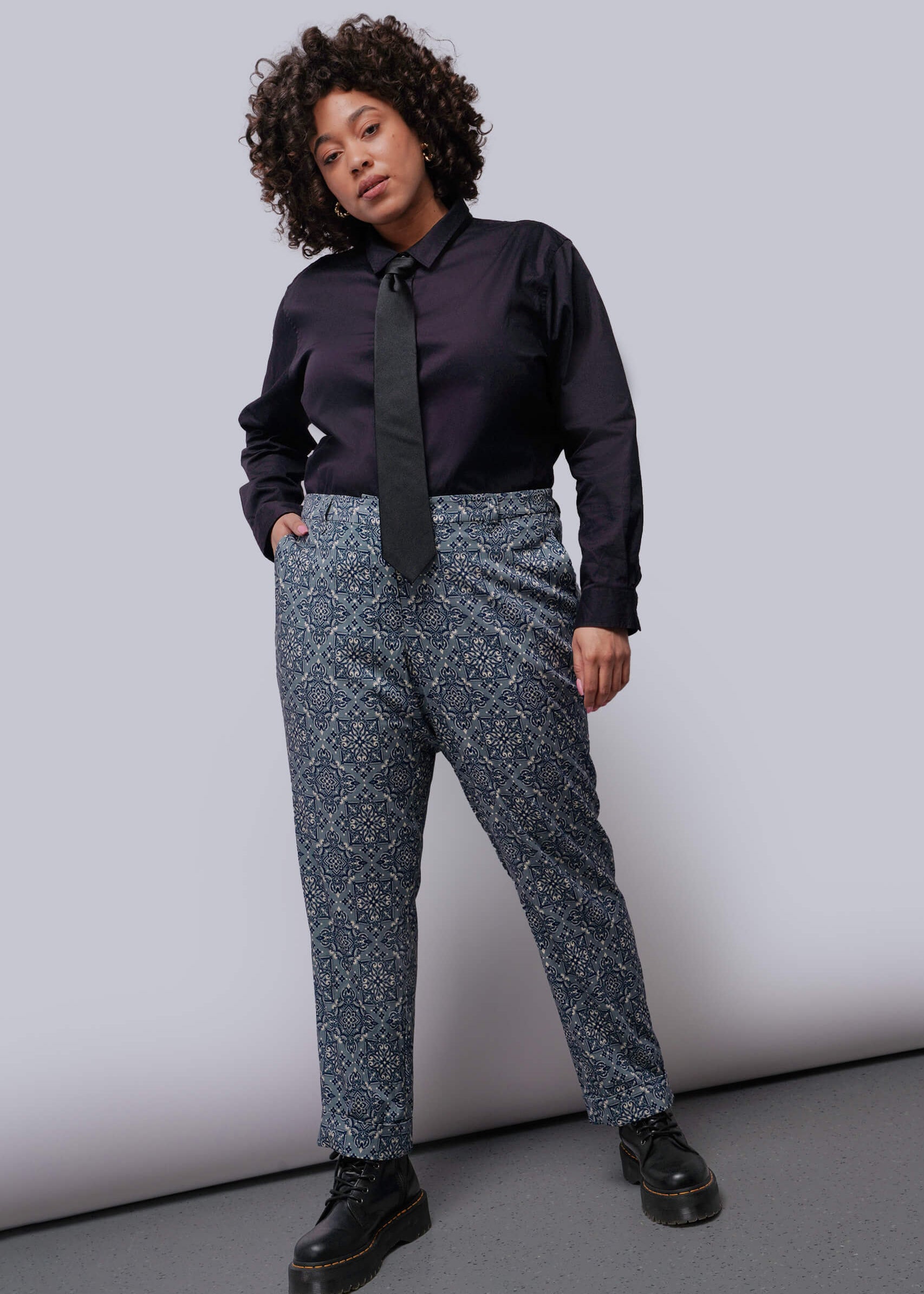 1X=Person wearing a black shirt, black tie, patterned pants, and black shoes against a plain background