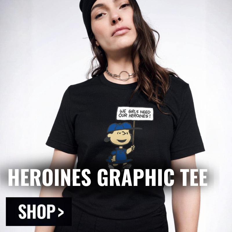 Person models the Peanuts x Wildfang Heroines Tee with text overlay in bottom left corner that reads HEROINES GRAPHIC TEE and button underneath that reads SHOP.