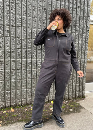 1X=Wearing The Essential Long Sleeve Coverall in an oversized fit, a person with short curly hair sips from a cup, standing against a textured gray wall. They sport black shoes while scattered leaves on the ground enhance the cozy autumn vibe.