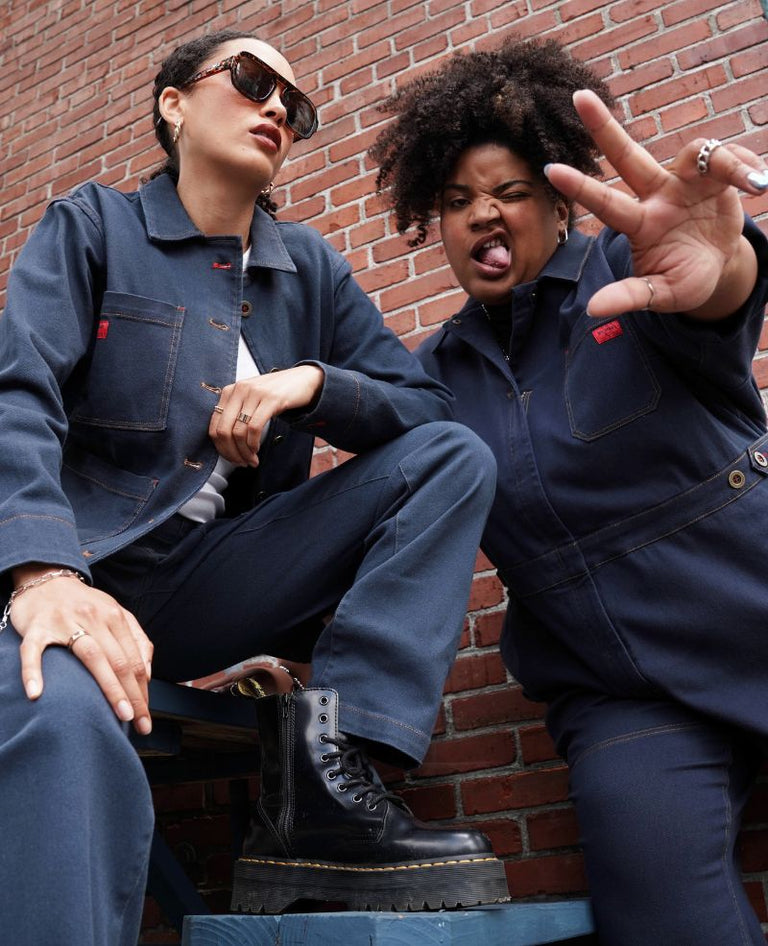 Two models wearing pieces from the Denim Collection including The Essential Denim Utility Jacket and Pant, and The Essential High Waisted Long Sleeve Denim Coverall in Navy Denim. They are crouching in front of brick wall outside