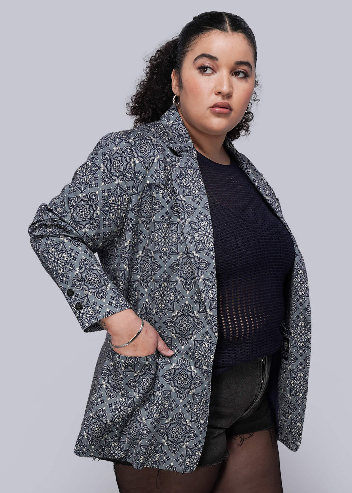 1X=Person wearing a patterned teal blazer over a navy blue mesh top, black shorts, and tights, standing against a plain background.