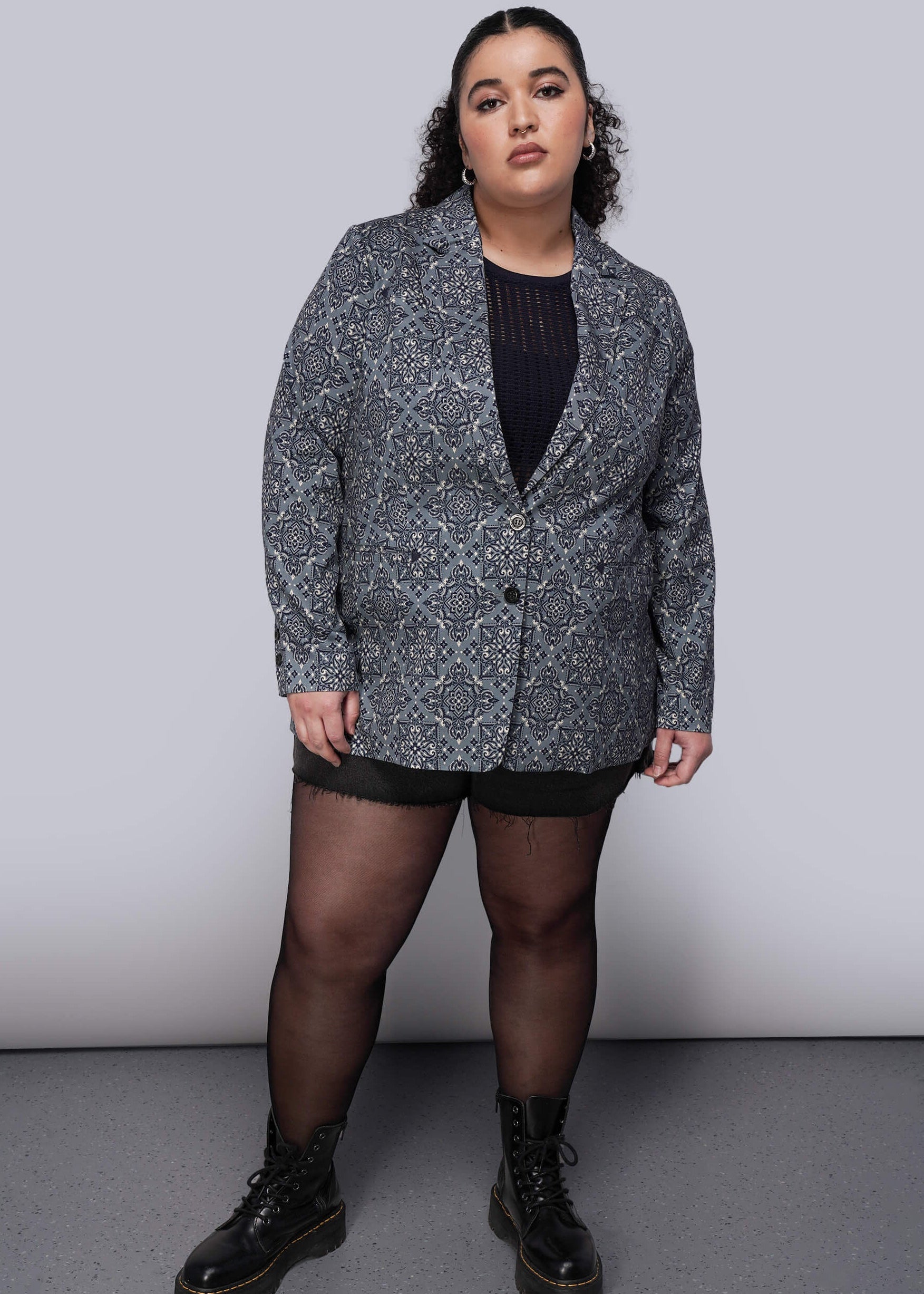 1X=Person wearing a patterned blazer over a black top with black tights and boots on a plain background