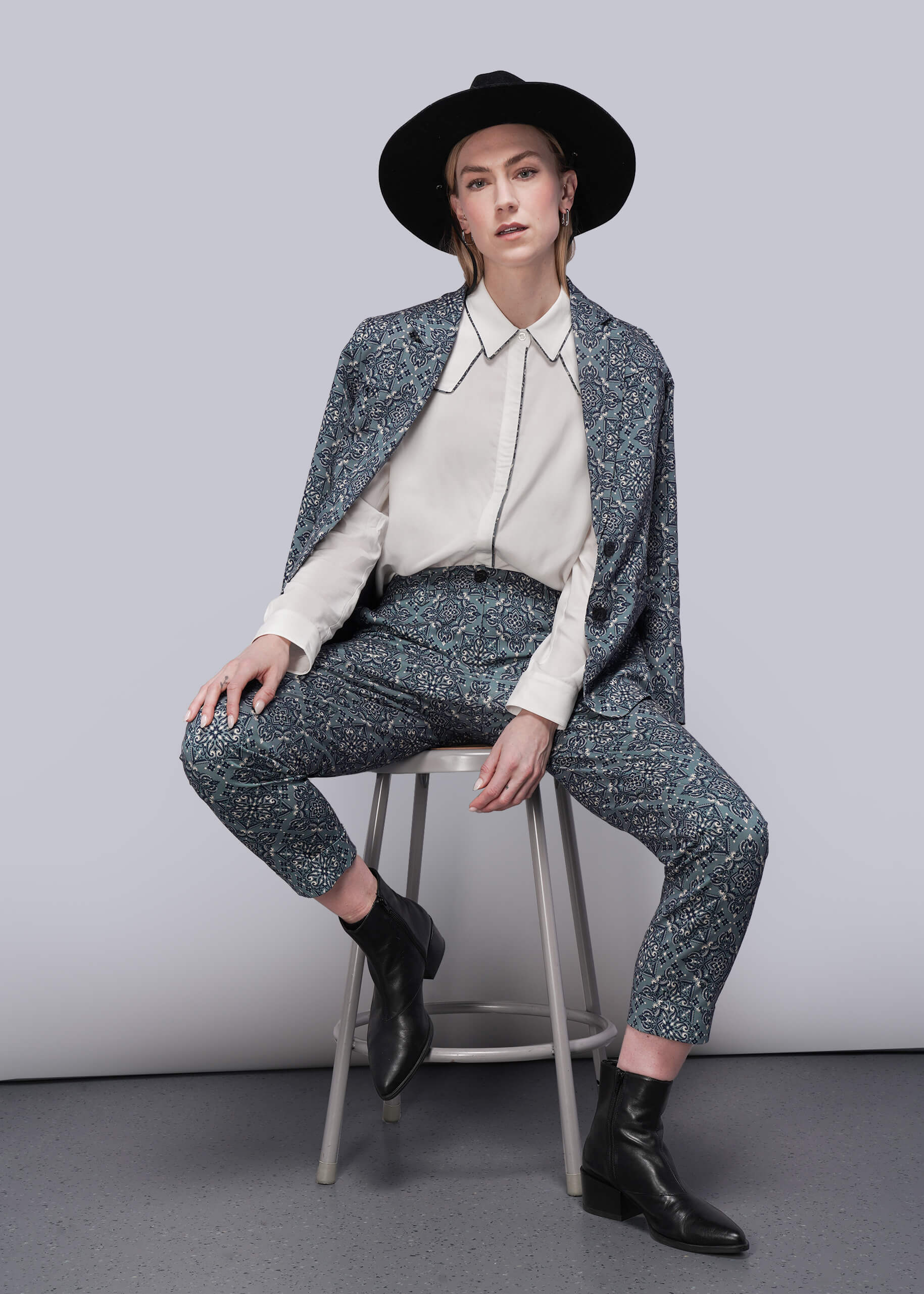 S=Person wearing a patterned suit and black hat sitting on a stool against a plain background