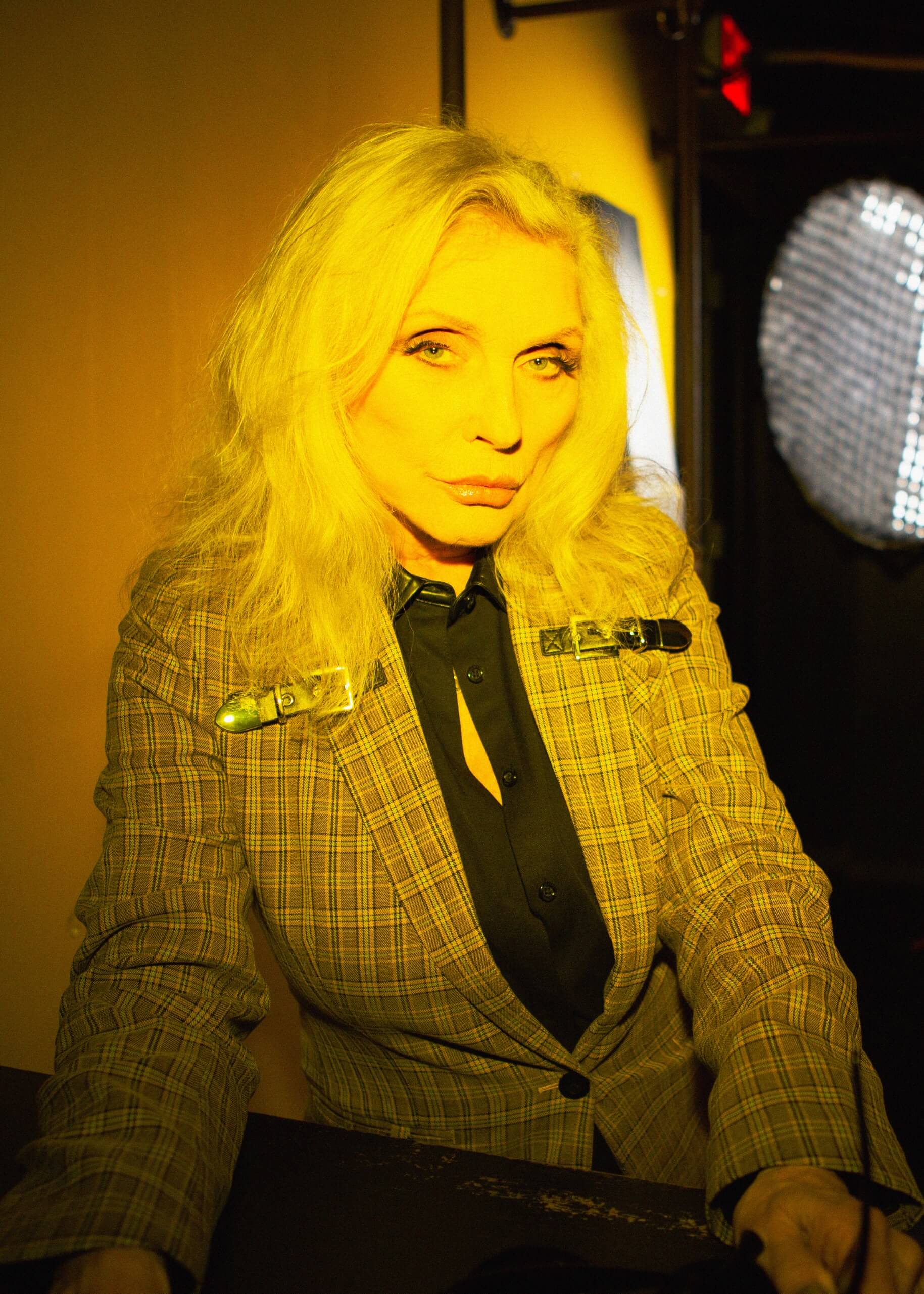 Person with blonde hair wearing a checkered suit in a dimly lit room.