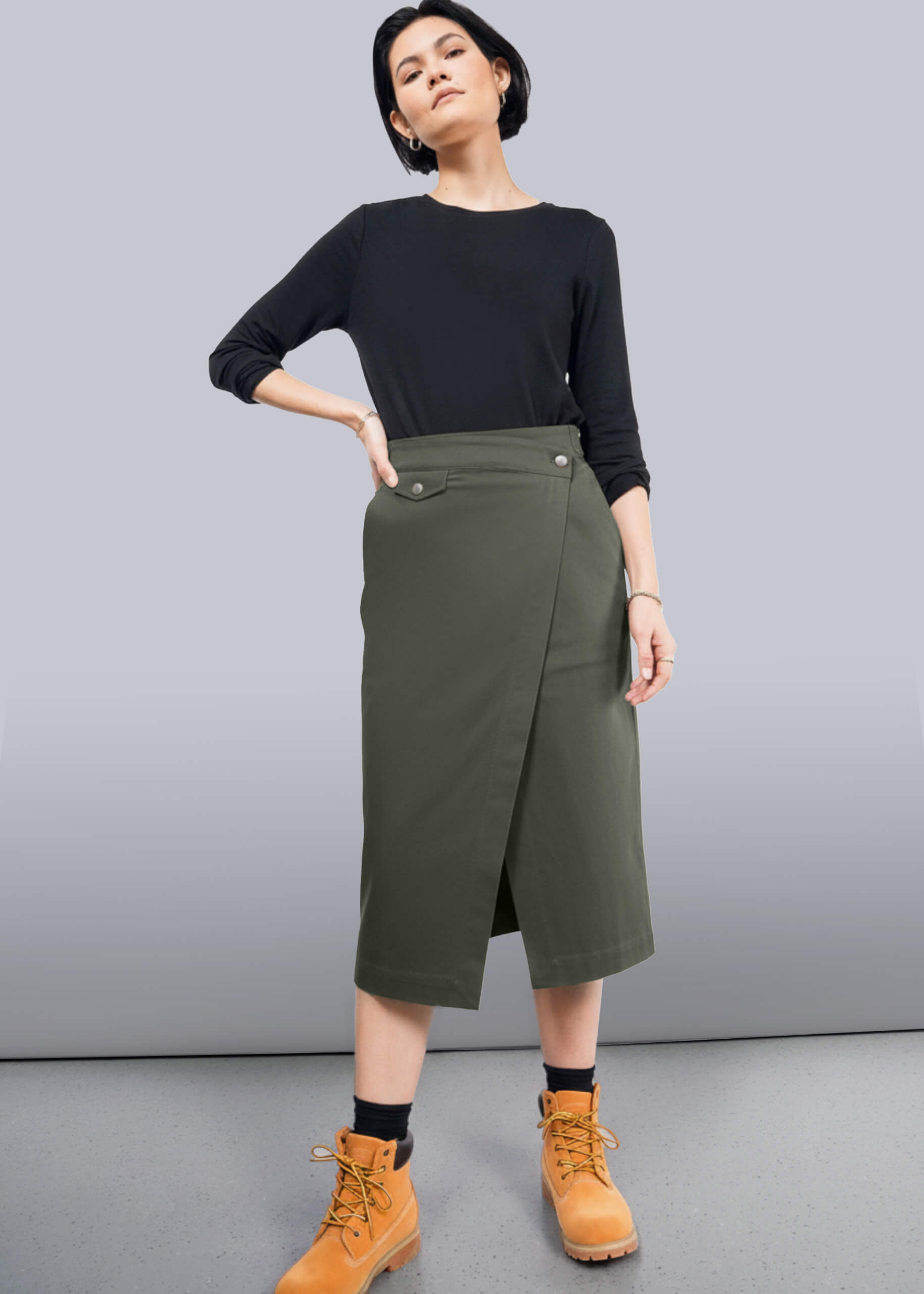 S=Woman wearing a black top and green skirt with orange boots on a gray background