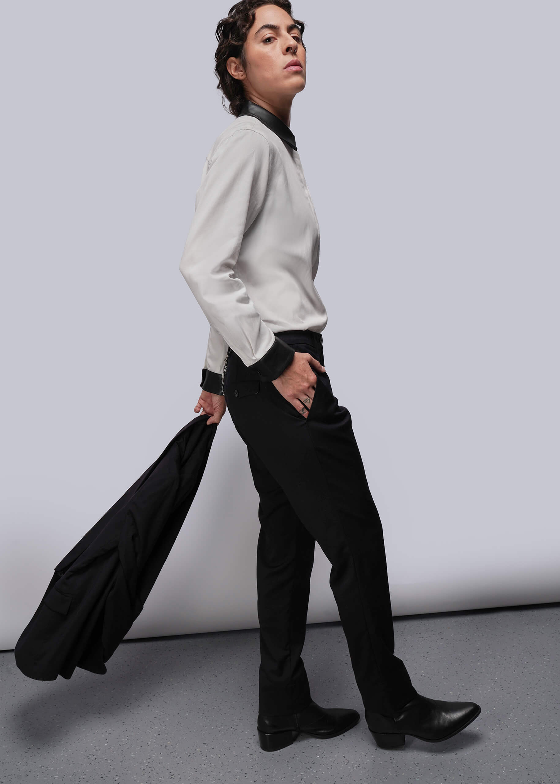 S=Person wearing a light-colored shirt and black pants holding a black blazer against a plain background