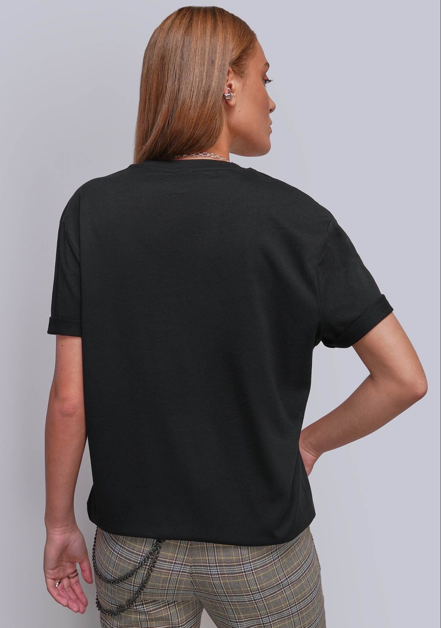 S=A woman shown from the back wearing a plain black short-sleeve t-shirt and beige plaid trousers, standing against a light gray studio background.