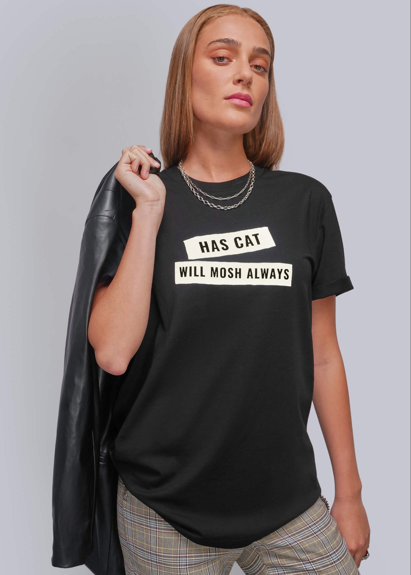 S=A woman modeling a black graphic t-shirt with the text 'HAS CAT WILL MOSH ALWAYS' in white block letters. She is wearing plaid pants and holding a black leather jacket over her shoulder, set against a plain gray studio background.