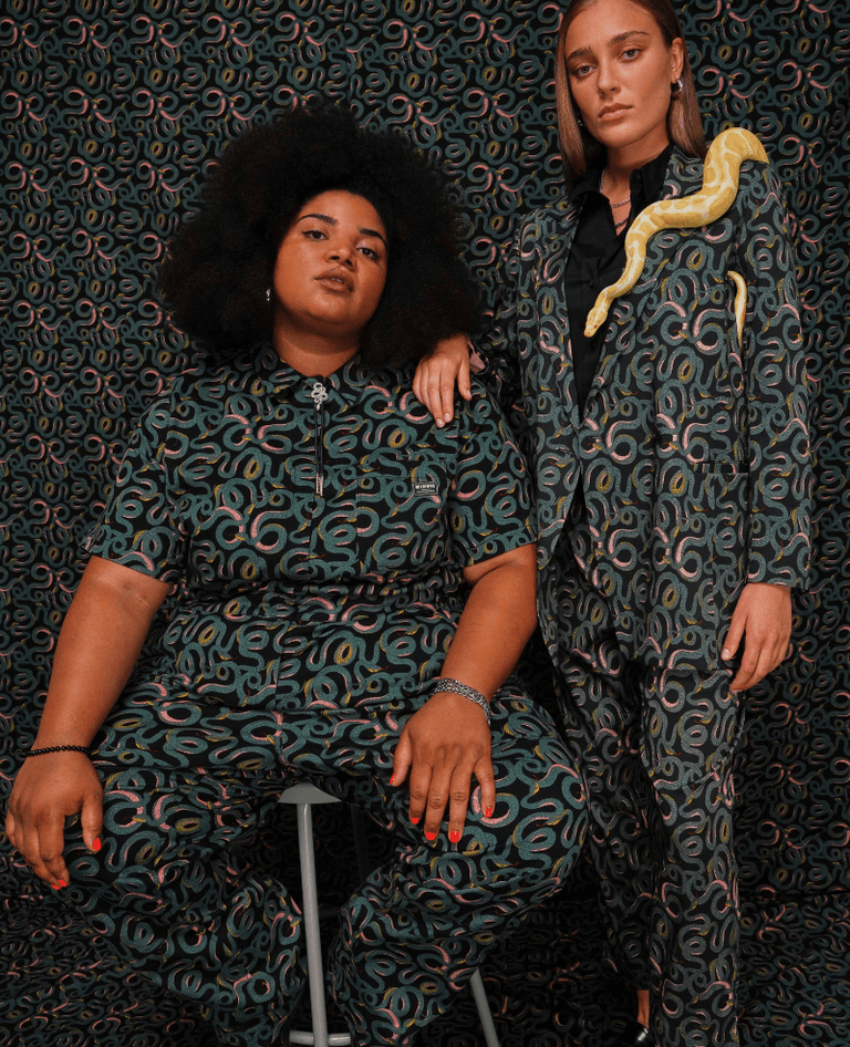 Against snake patterned background, two models wear The Essential High Waisted Coverall, The Empower Double Breasted Blazer, and The Empower Slim Crop Pant all in Serpentine Black, which is a snake print.