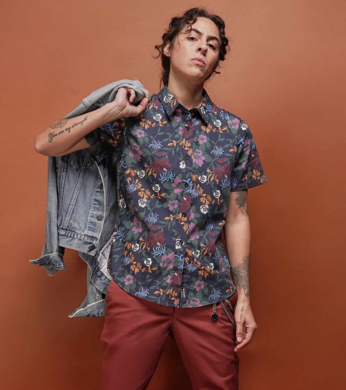 Against orange background, person wearing floral button up and red pants holds jean jacket over shoulder while looking at camera.