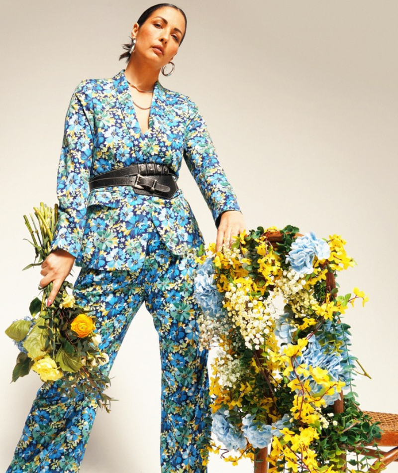 Person wearing The Empower Tux Blazer and The Empower Wide Leg Pant in Carnations Navy, which is a blue toned floral print, looks at camera holding a yellow toned bouquet. One hand rests on a chair covered in yellow, blue, and white flowers.