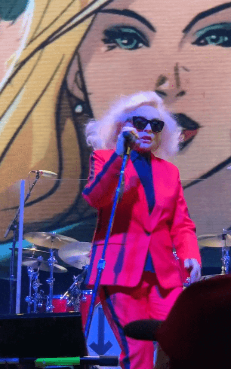 Debbie Harry in a red suit performing on stage with musical instruments and a large screen in the background.