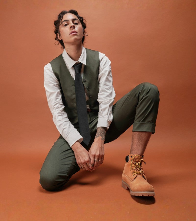 Against orange background, person kneels while wearing green vest, blazer, and a long sleeve white button up.