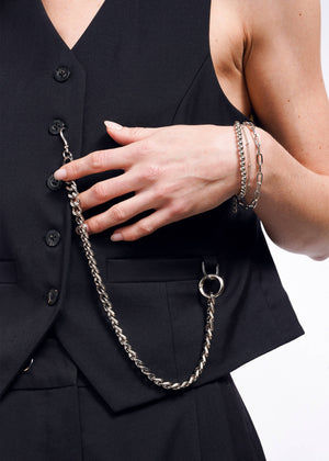 S= A person wearing the Debbie Harry X Wildfang Vest, with a large silver chain hanging from a pocket. Their hand rests on the vest, adorned with silver bracelets, highlighting a unique blend of fashion and jewelry.