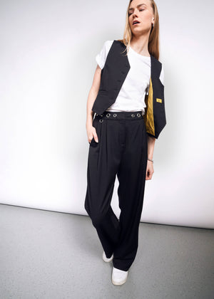 S=Person posing casually against a white background, wearing a white T-shirt, black open vest, Debbie Harry X Wildfang Wide Leg Trousers, and white sneakers. Their head is tilted slightly and one hand is in their pocket.