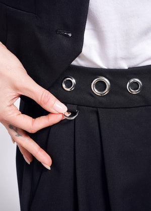 S=A person in Debbie Harry X Wildfang wide leg trousers with large silver grommets adjusts a waistband detail. A finger tattoo peeks out as they stylishly pair it with a crisp white shirt, completing the look.