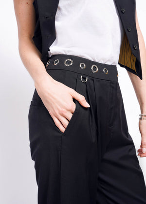 S=Someone poses with a hand in the pocket, wearing sleek black Debbie Harry X Wildfang Wide Leg Trousers with a grommet waistband, a black vest, and a white shirt. The outfit captures Debbie Harrys edgy style through its modern design and striking metal accents.