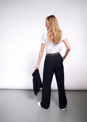 S=A woman with long hair, wearing a white t-shirt and Debbie Harry X Wildfang Wide Leg Black Trousers featuring grommet details, stands facing away holding a black jacket. The background is plain, and the floor is light gray.
