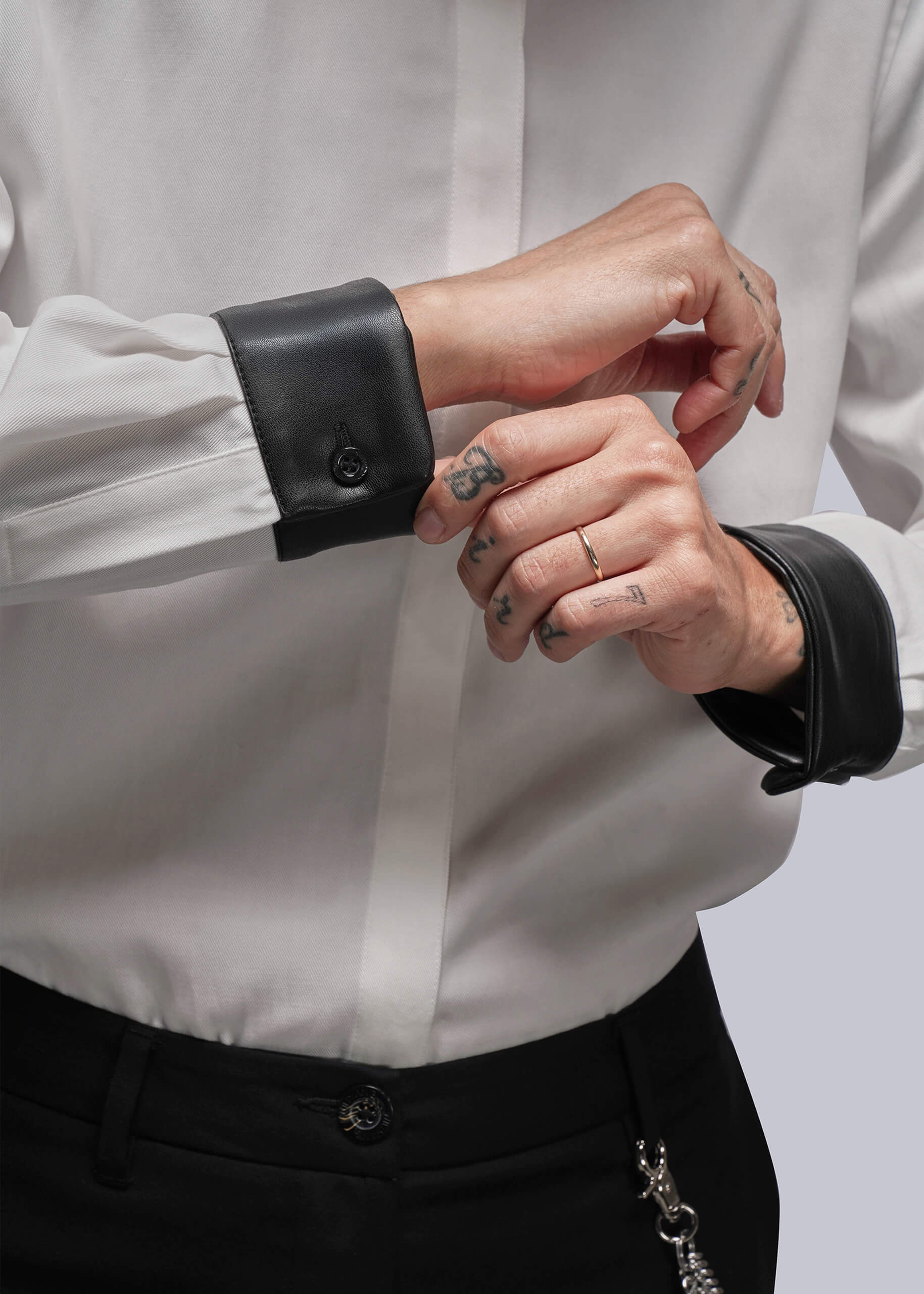 S=Close-up of a person wearing a white shirt with black cuffs and black pants on a light gray background.