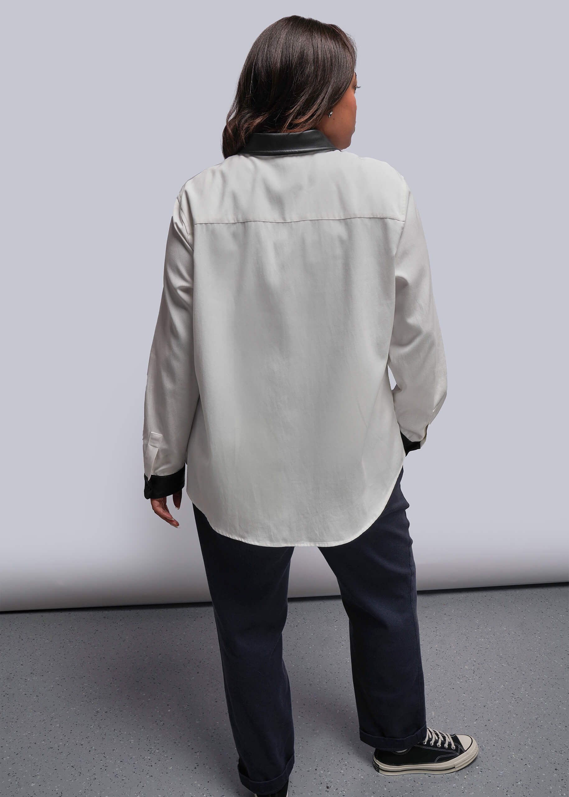 1X= Back view of person wearing a white button up with vegan leather cuffs and collar and dark pants against a plain background