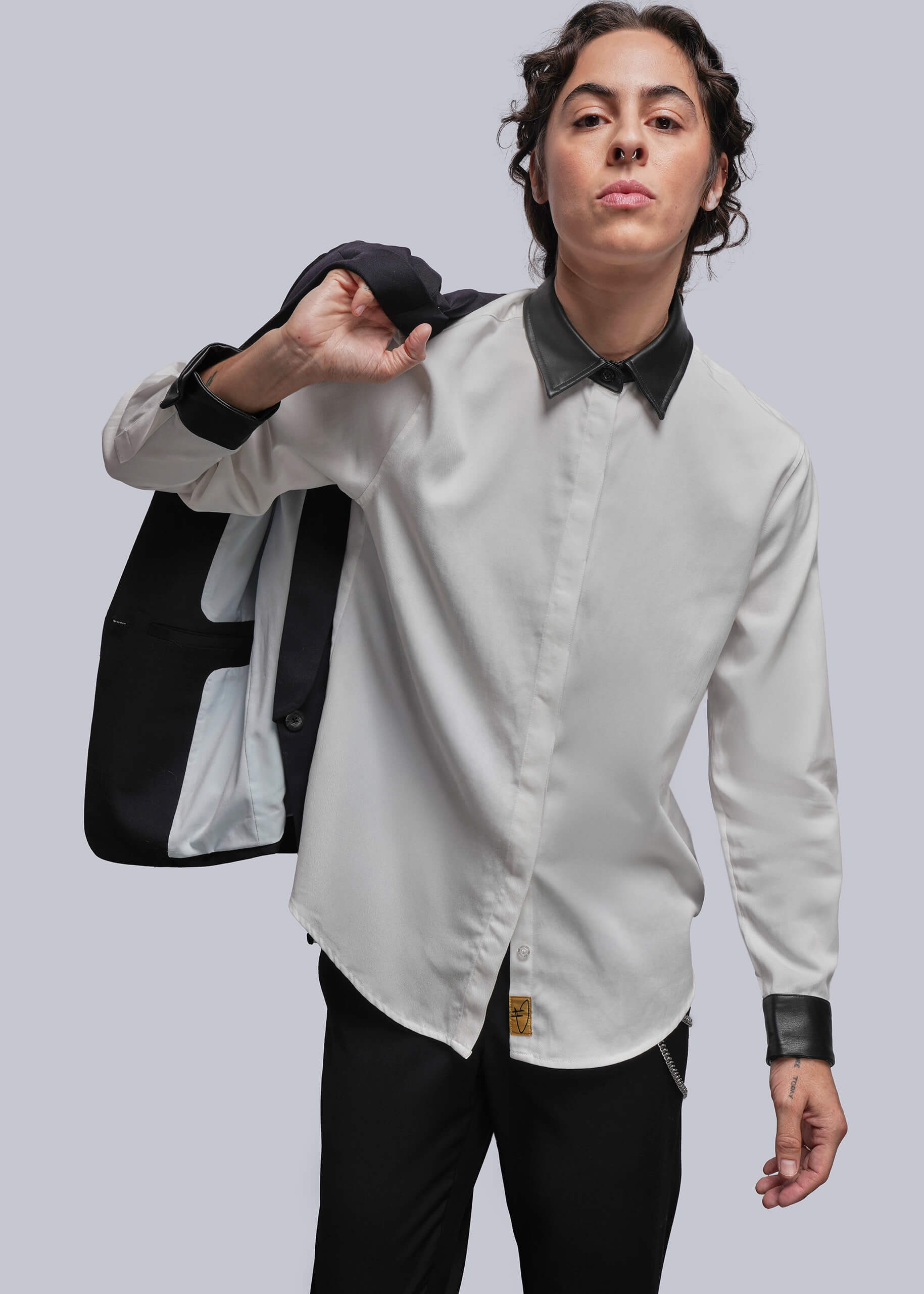Person wearing a white shirt with black collar and cuffs, holding a black blazer against a gray background