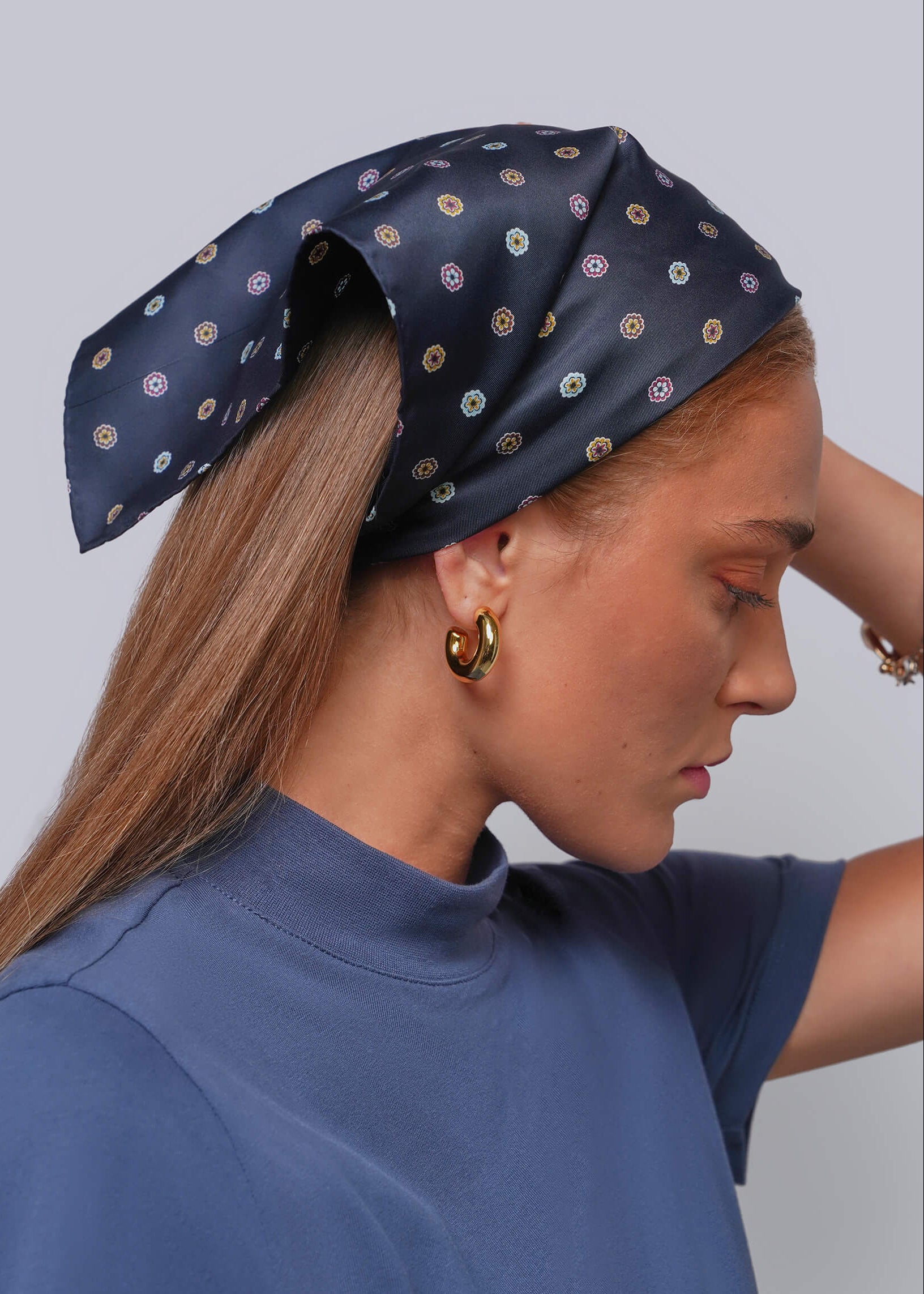 A woman in profile wearing a navy blue silk-like headscarf with a small multicolor circular pattern, paired with a blue mock-neck top and gold hoop earrings, against a solid gray background.