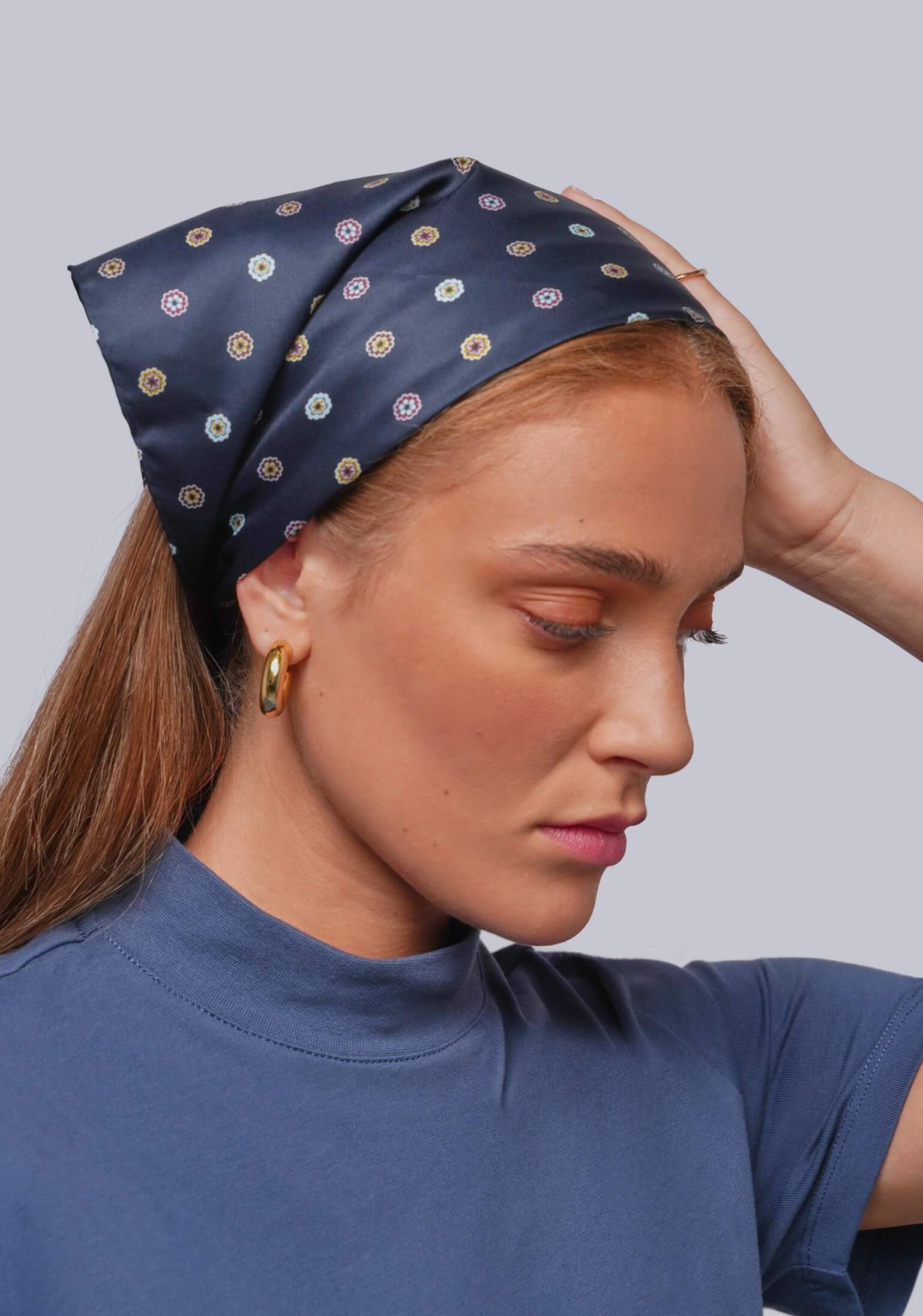 A woman wearing a navy blue silk-like headscarf with a repeating circular pattern in beige, white, and muted red, paired with a blue t-shirt against a solid gray background.