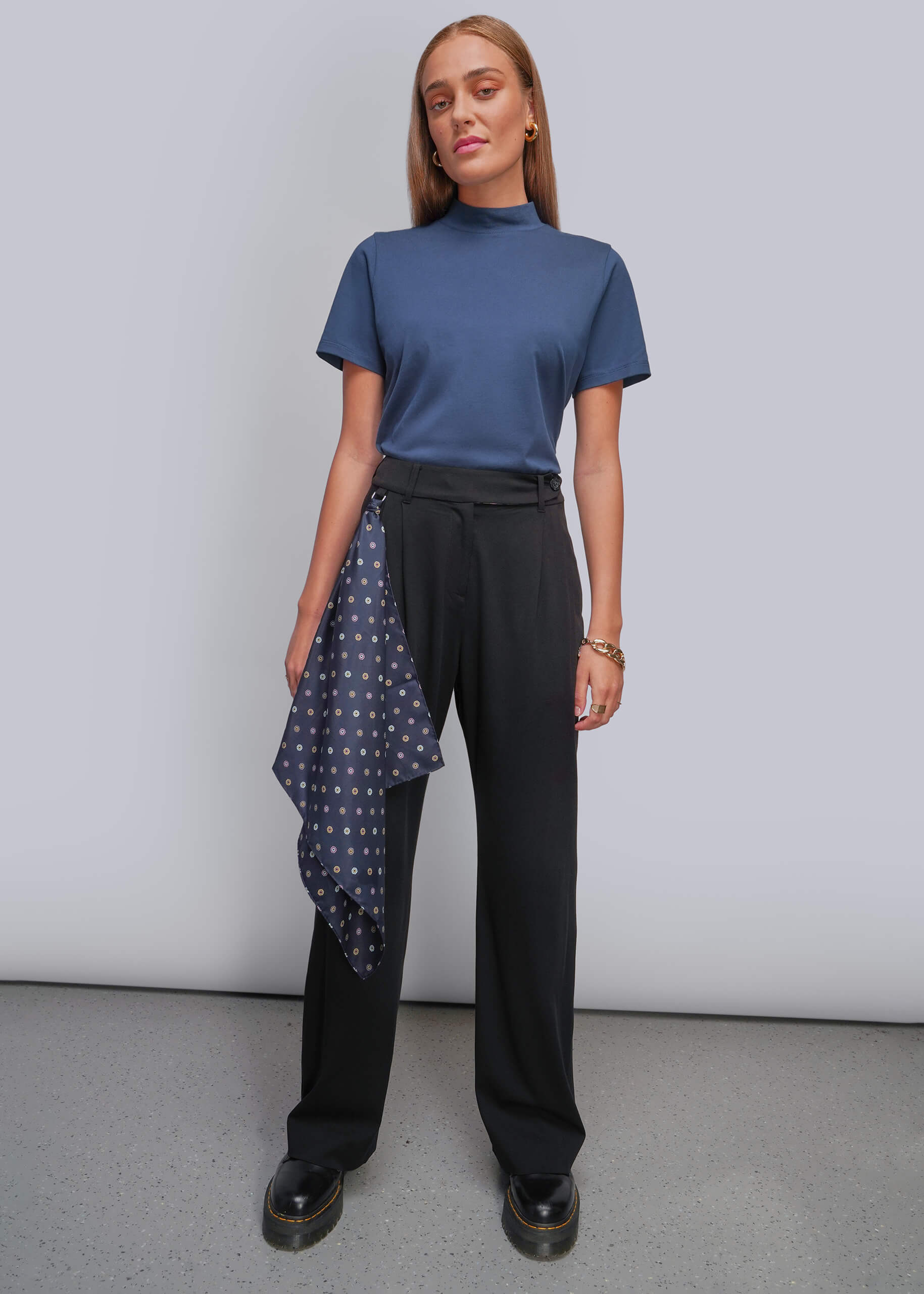 A woman modeling a slate blue mock neck short-sleeve top, black wide-leg trousers, and a navy blue polka dot scarf hanging from their pants, standing against a light gray studio background.