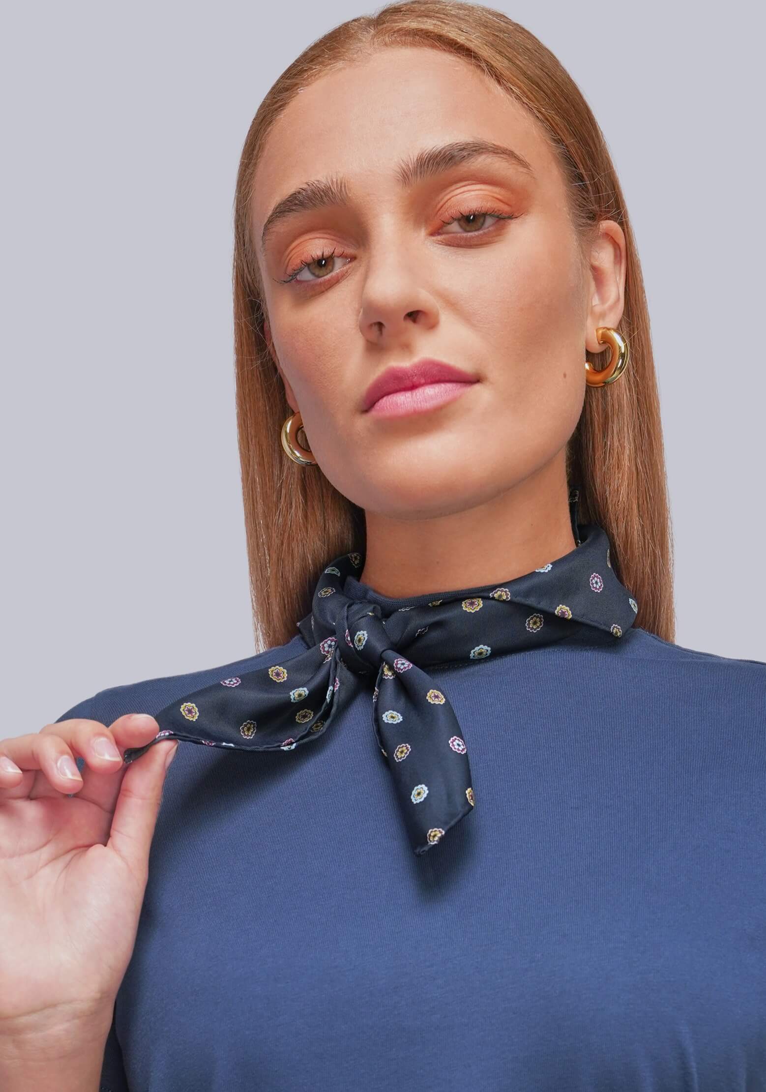 A woman wearing a navy blue silk-like neck scarf with a small gold and white pattern, tied in a knot at the collar of a navy top, against a solid gray background.