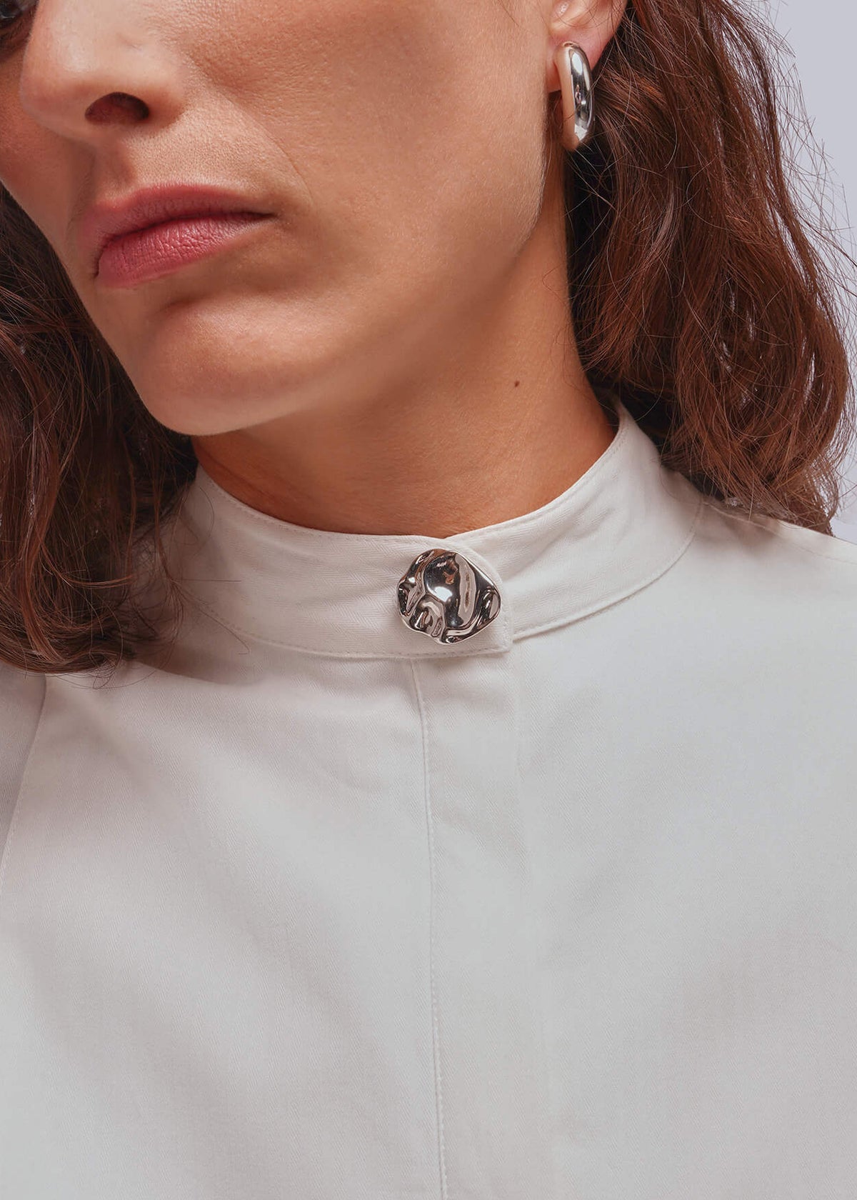 A close-up of a woman wearing a white high-neck blouse with a decorative silver-toned button cover at the collar, against a light gray background.