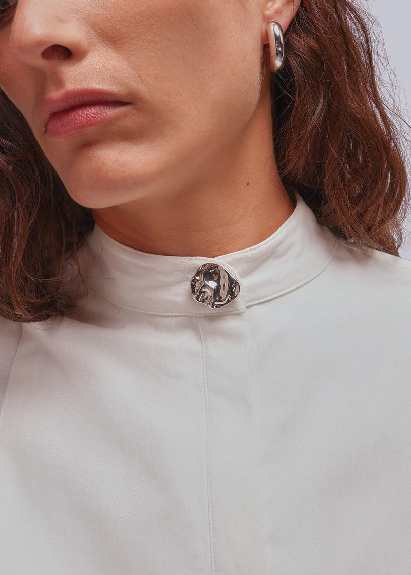 A close-up of a woman wearing a white high-neck blouse with a decorative silver-toned button cover at the collar, against a light gray background.