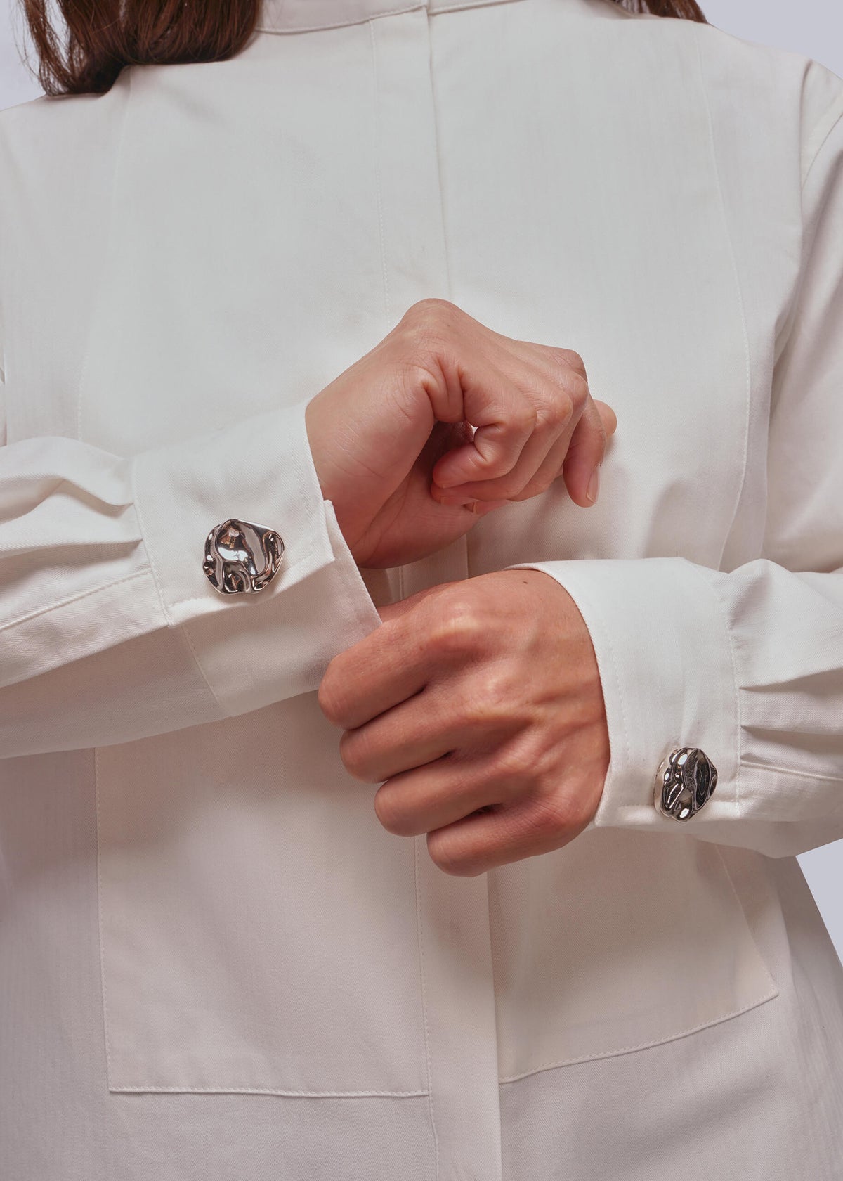 Close-up of a person wearing a white long-sleeve shirt or coat, adjusting the cuff with one hand while the other hand rests near the cuff. The shirt has decorative round silver-toned button covers and a clean, structured silhouette.