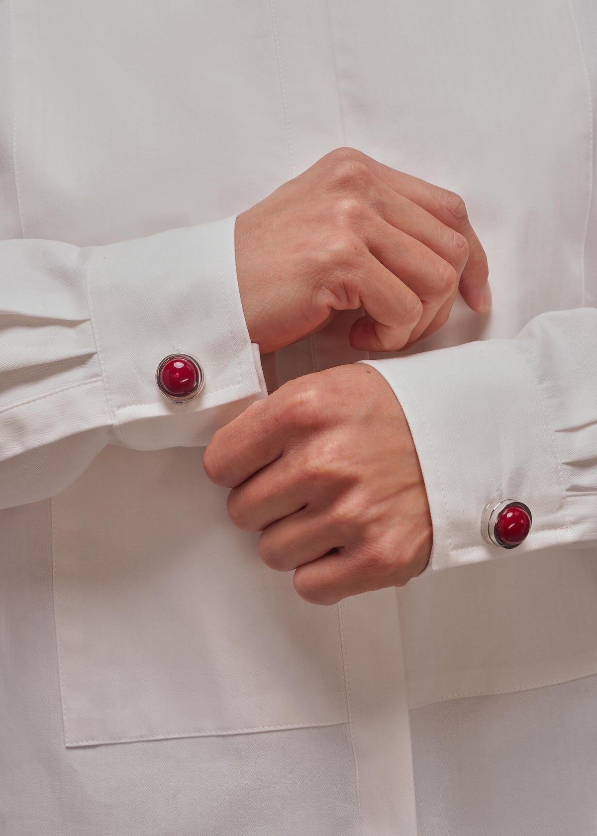 Close-up of a person's hands adjusting the cuffs of a white long-sleeve shirt, featuring decorative round red button covers with silver-toned metal rims.