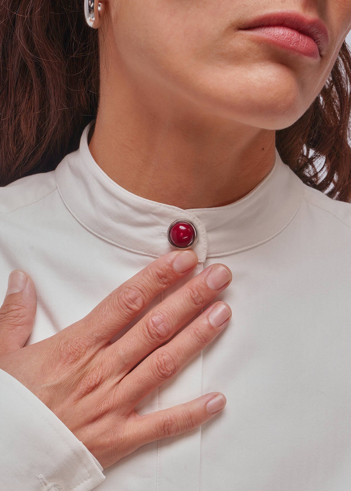 Close-up of a woman wearing a cream-colored high-neck blouse with a single red button at the collar, with her hand resting on her chest.