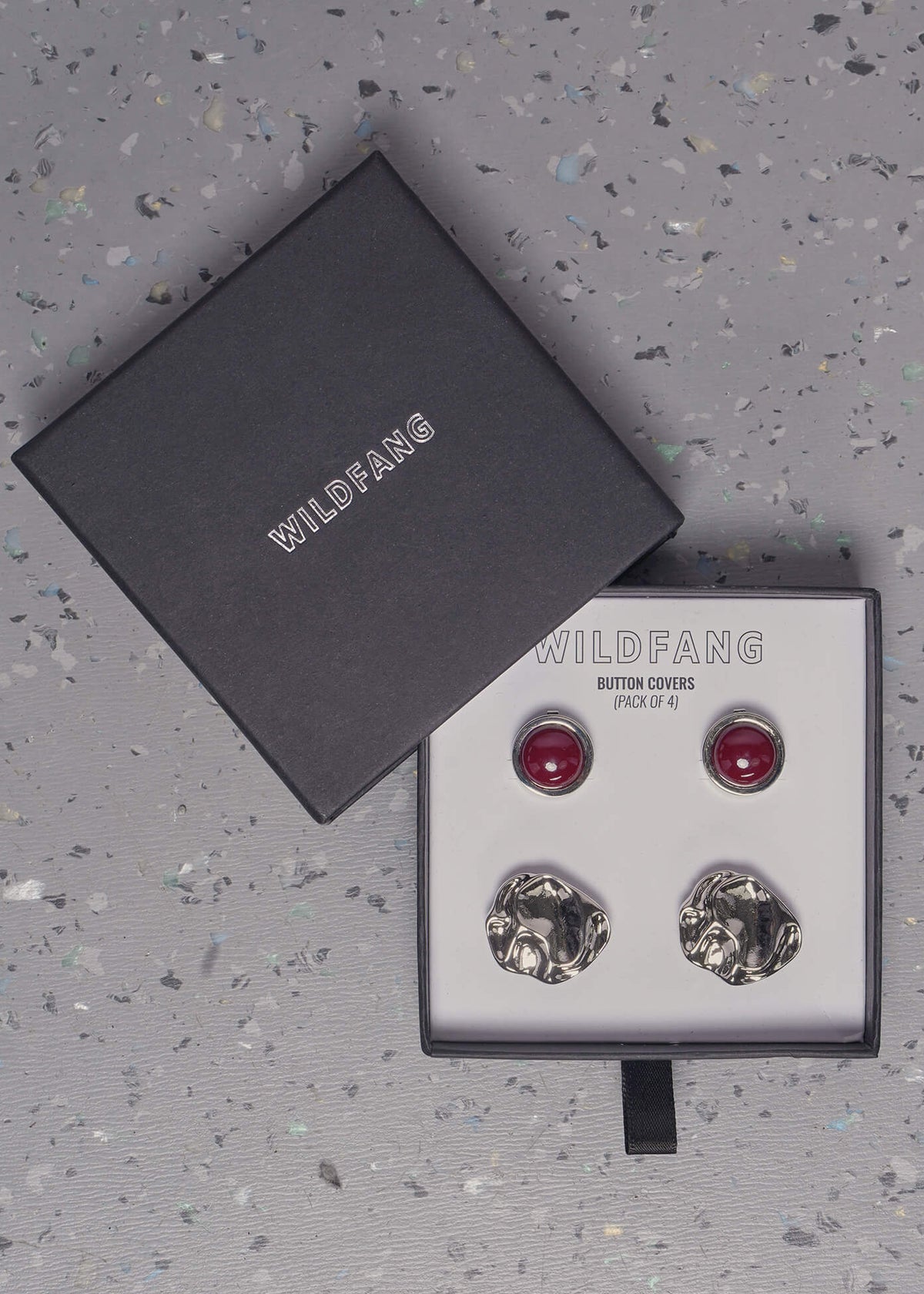A pair of Wildfang button covers displayed in their original black gift box on a speckled gray surface. The button covers feature two red round and two textured silver designs.