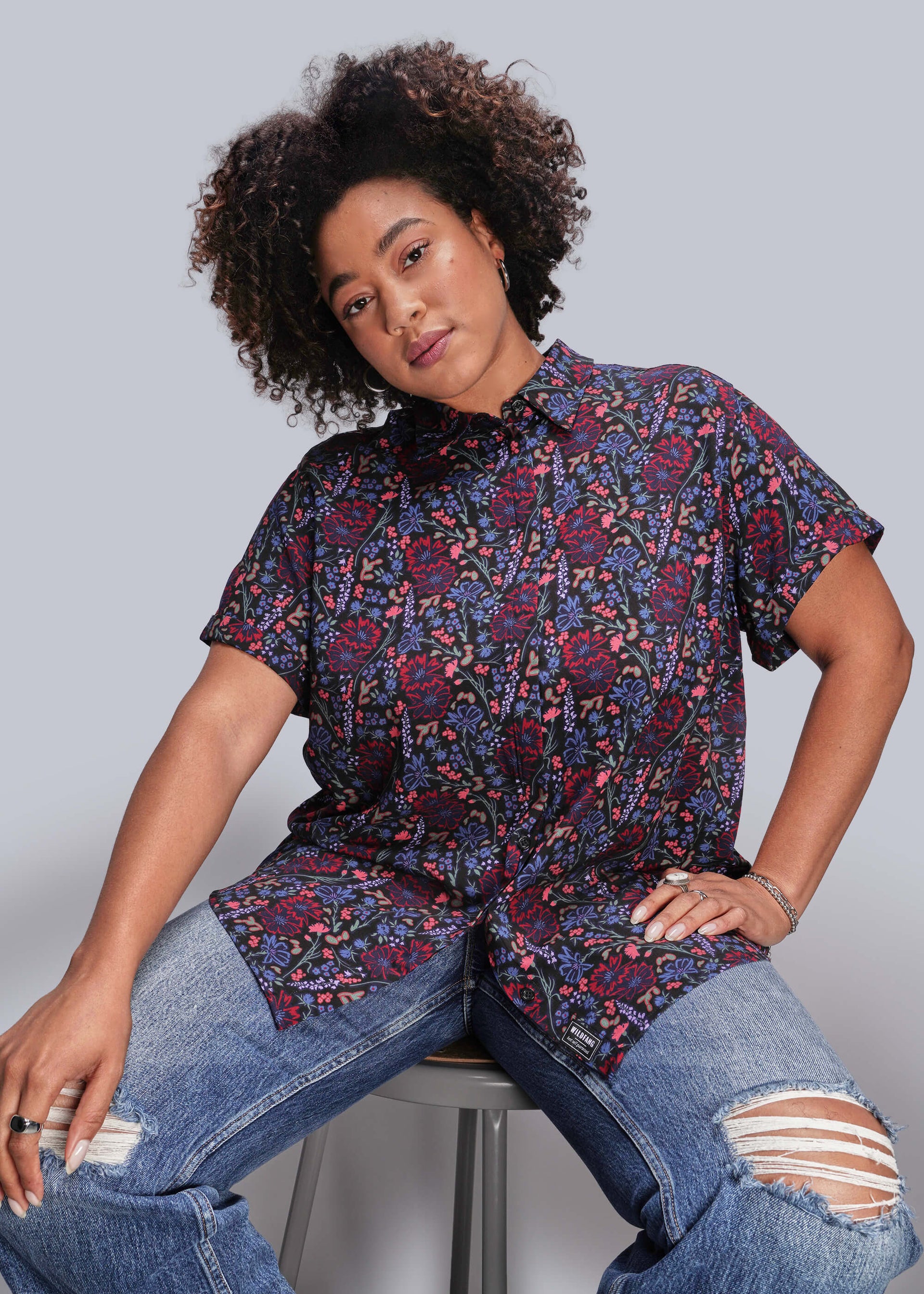 1X=Person wearing a patterned shirt and jeans sitting on a stool against a plain background