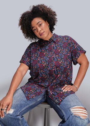1X=Person wearing a patterned shirt and jeans sitting on a stool against a plain background