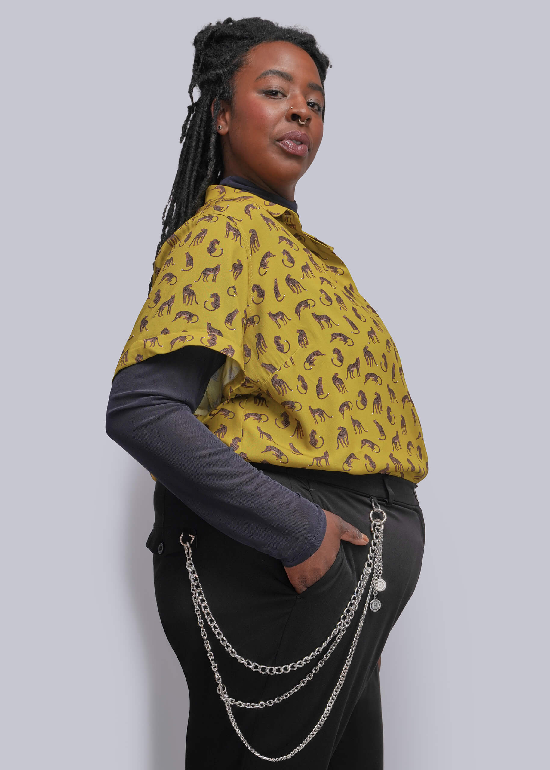 1X=A woman modeling a mustard yellow short-sleeve button-up shirt with a brown paisley pattern, worn over a dark gray long-sleeve top and black pants with a silver chain accessory, against a solid gray background.