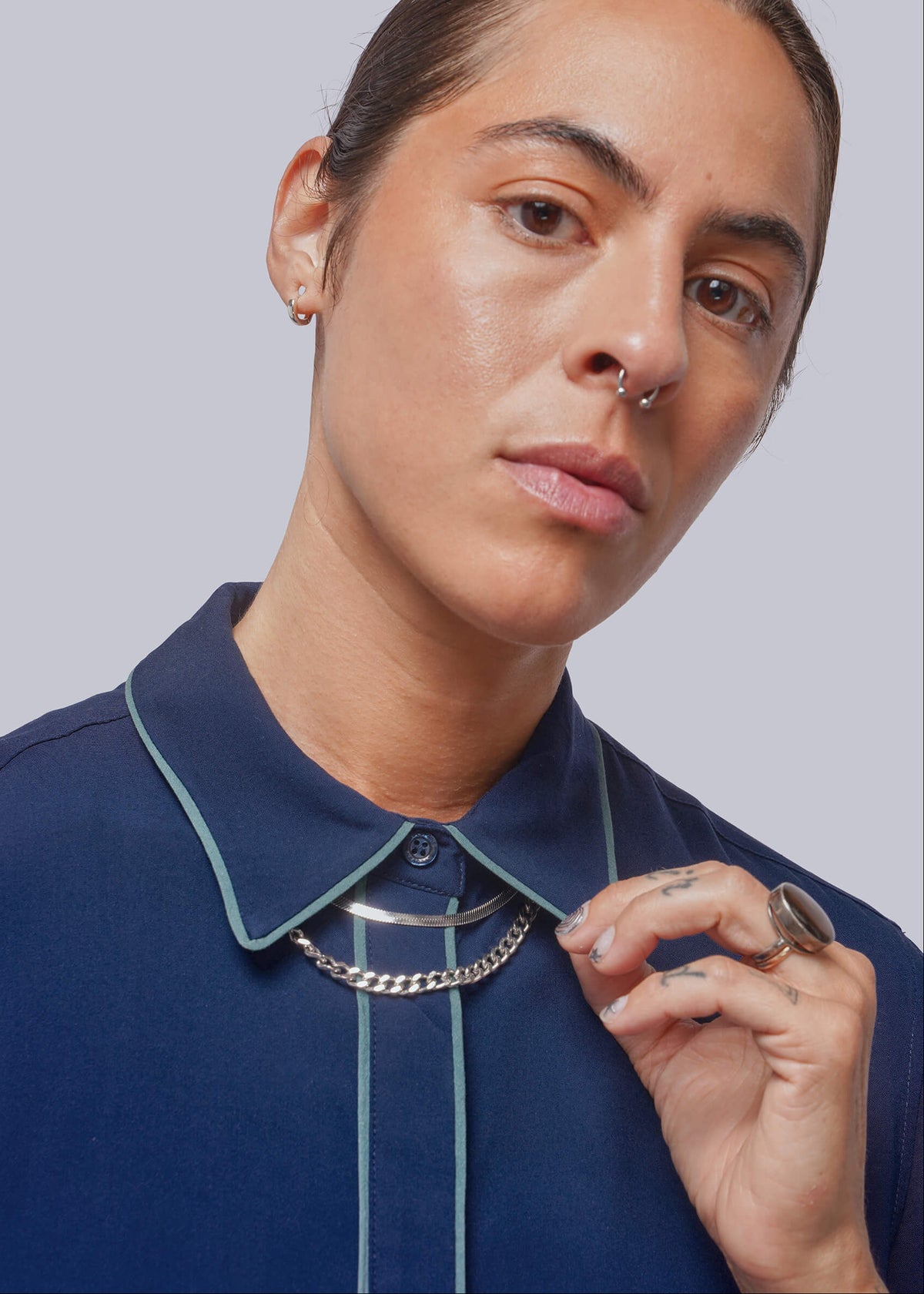S=A close-up of a model wearing a navy blue collared shirt with light green piping and a chunky silver chain necklace, set against a solid gray background.