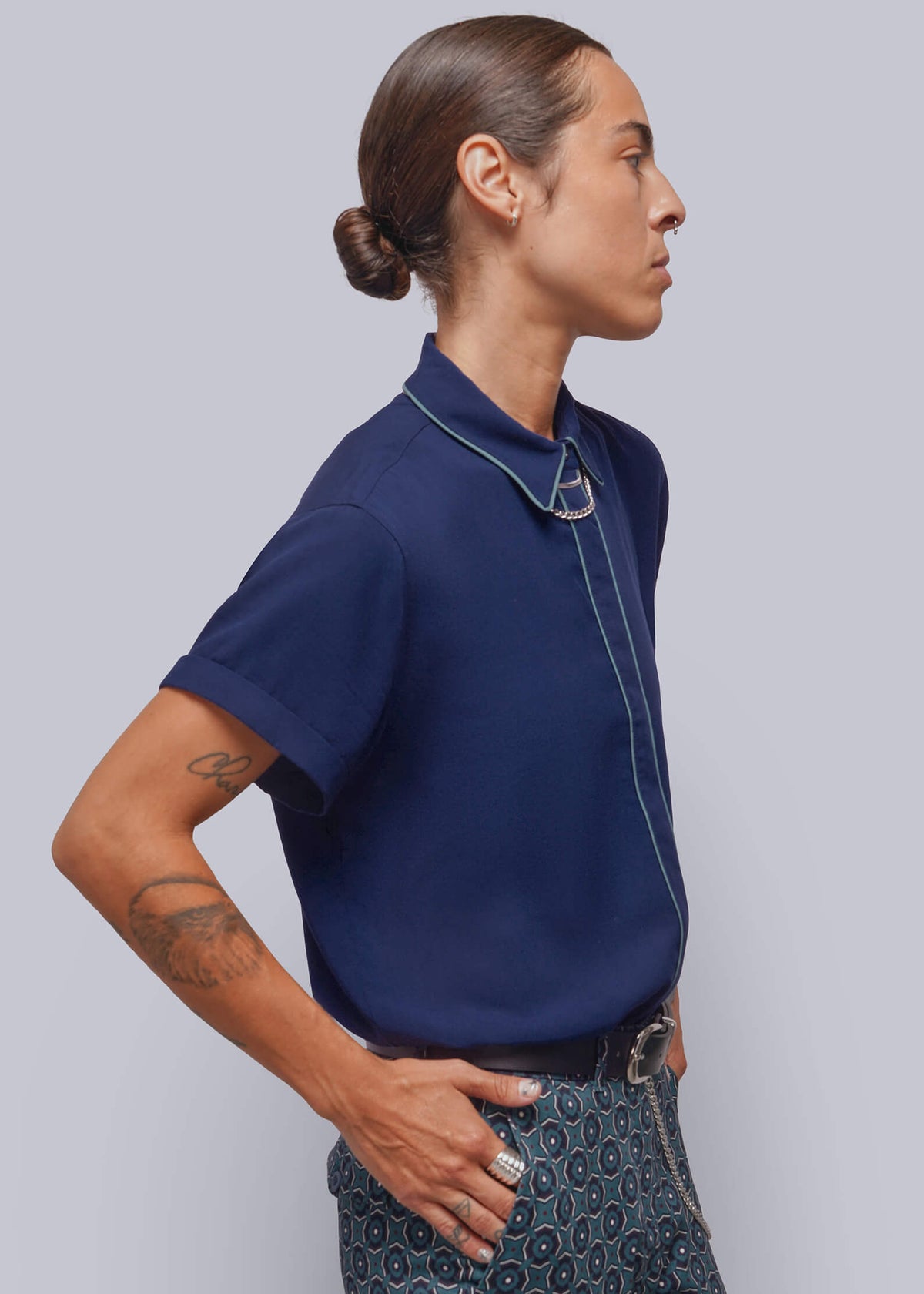 S=A model shown in profile wearing a navy blue short-sleeve polo shirt with light blue piping details and patterned trousers, set against a solid light gray background.