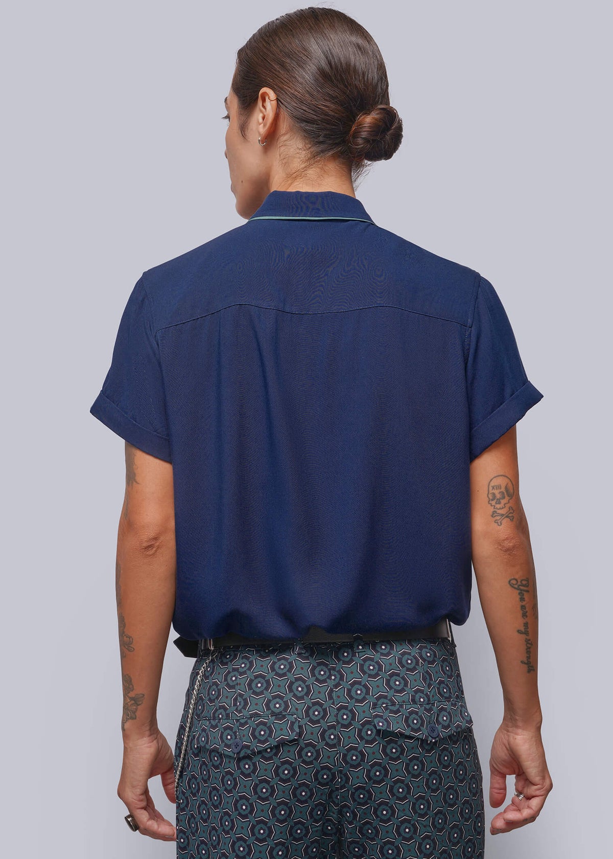 S=A model shown from the back wearing a navy blue short-sleeve button-up shirt and dark patterned trousers, set against a solid light gray background.