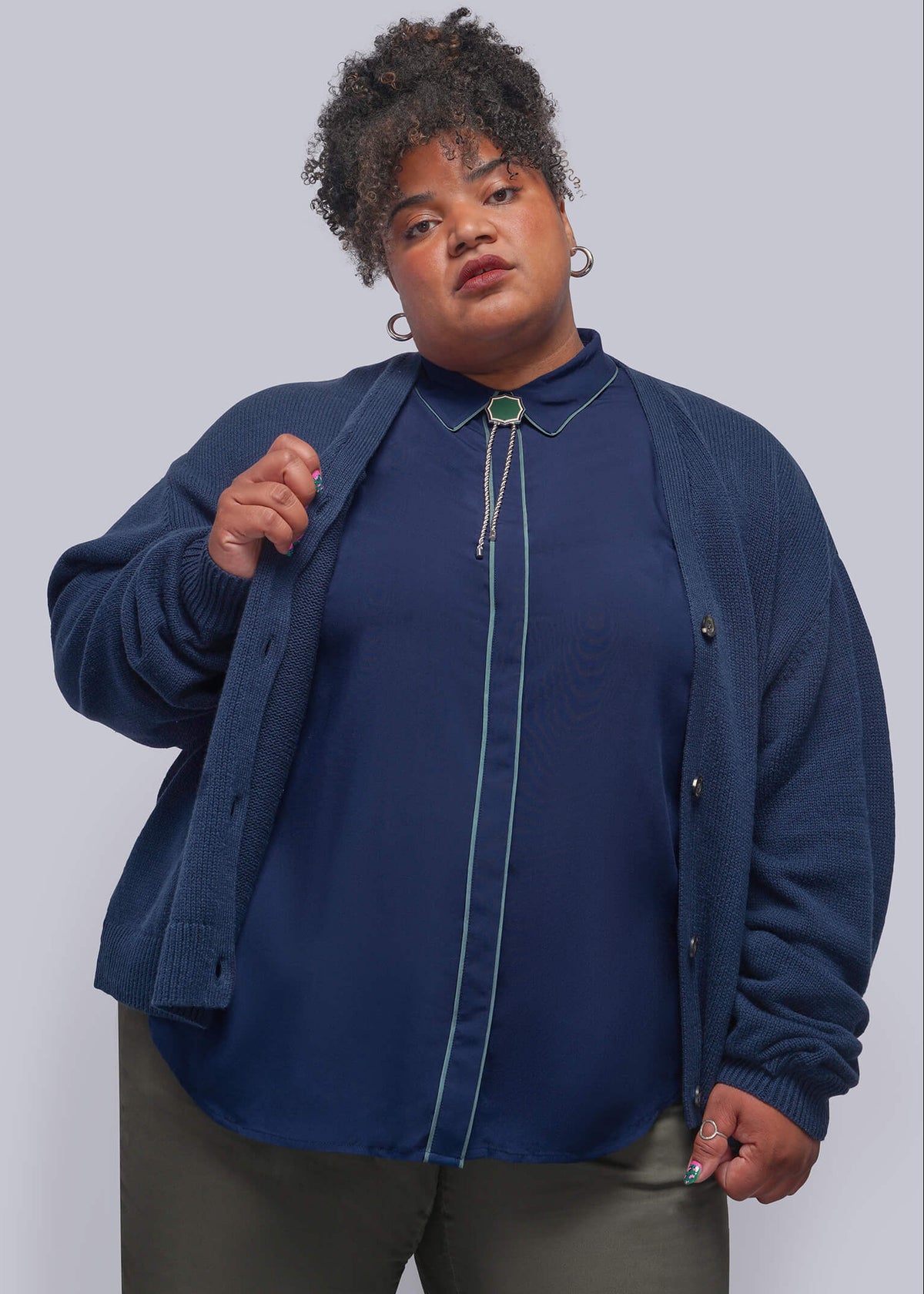 1X=A plus-size model wearing a navy blue zip-up cardigan over a matching navy top, styled with olive green pants against a solid light gray background.
