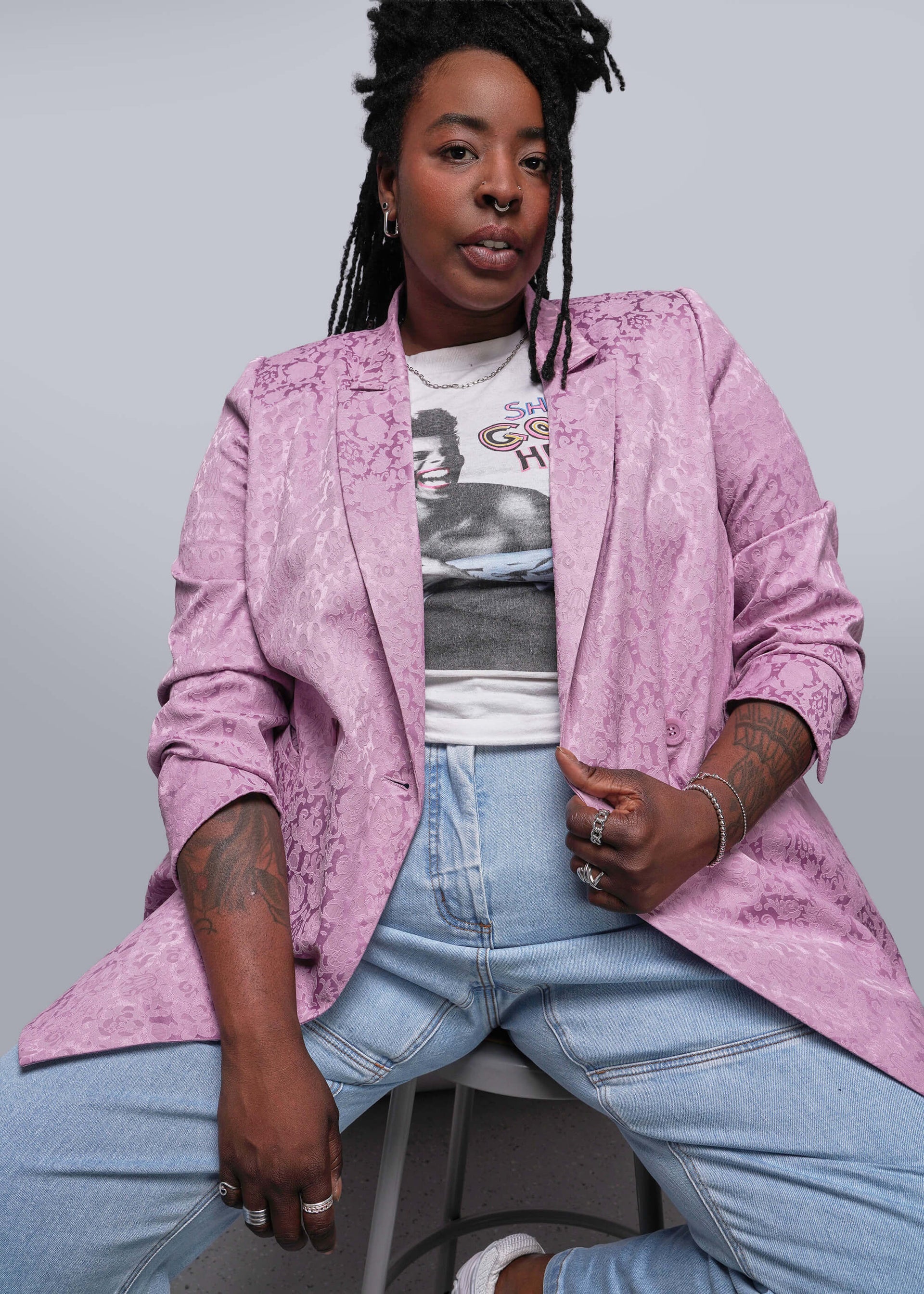 1X=Person wearing a pink jacket over a graphic t-shirt and blue jeans on a gray background