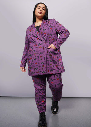 1X=Person with long black hair wearing purple bug printed double breasted blazer, matching slim crop pant, and Doc Marten boots looks at camera mid step.
