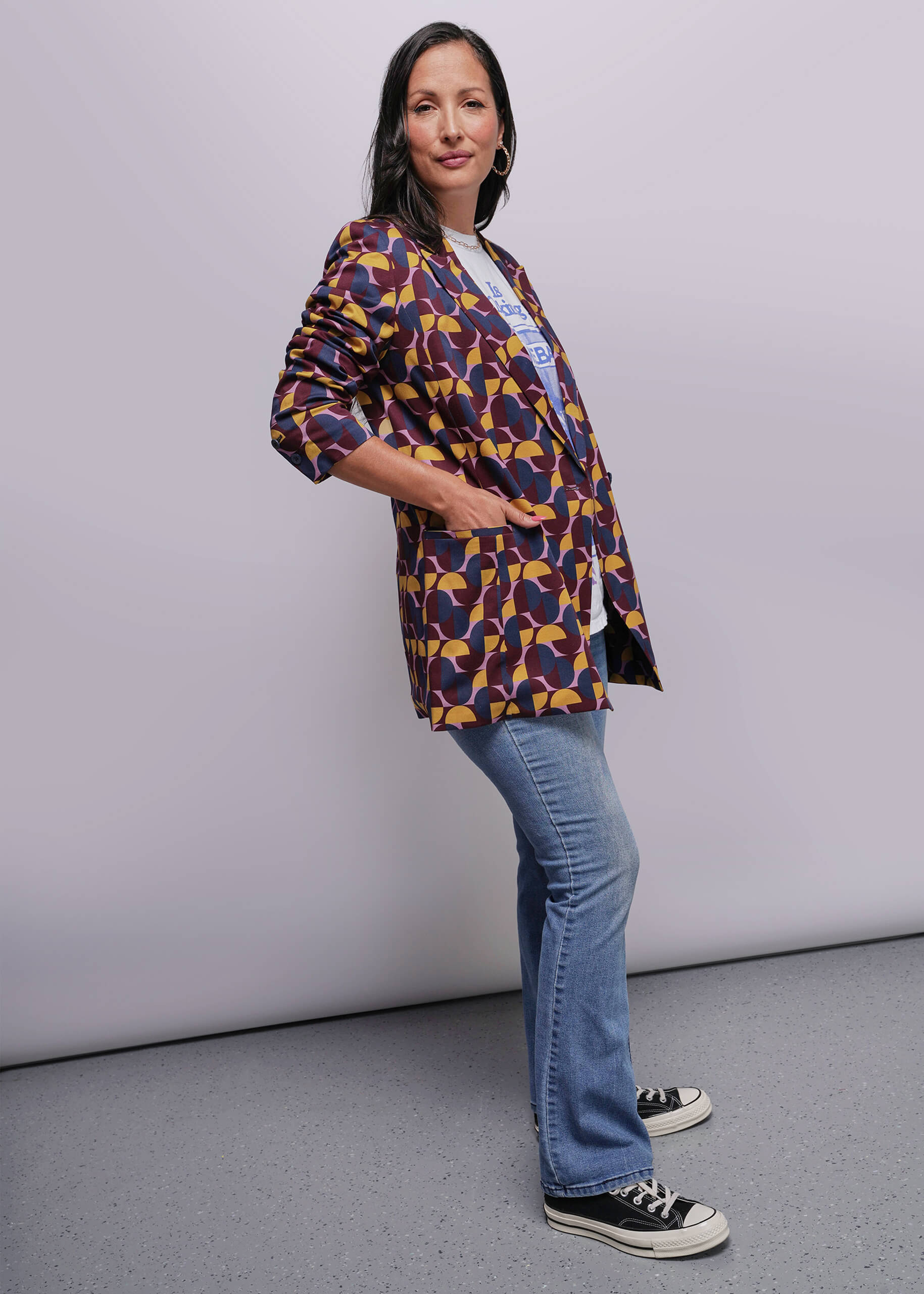 S=Woman wearing a colorful patterned jacket and blue jeans against a plain background