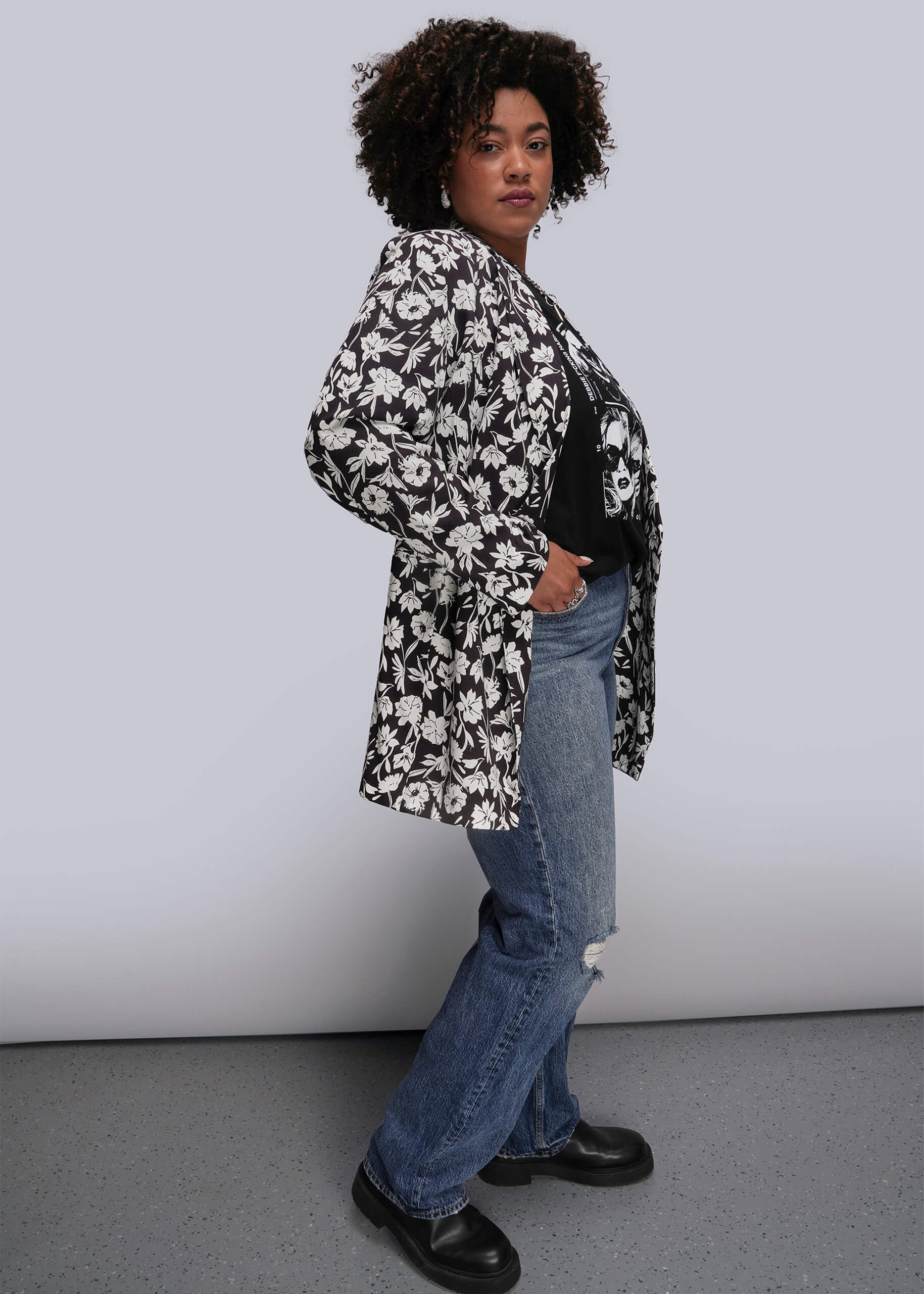 1X=Person wearing a black and white floral blazer with blue jeans against a plain background